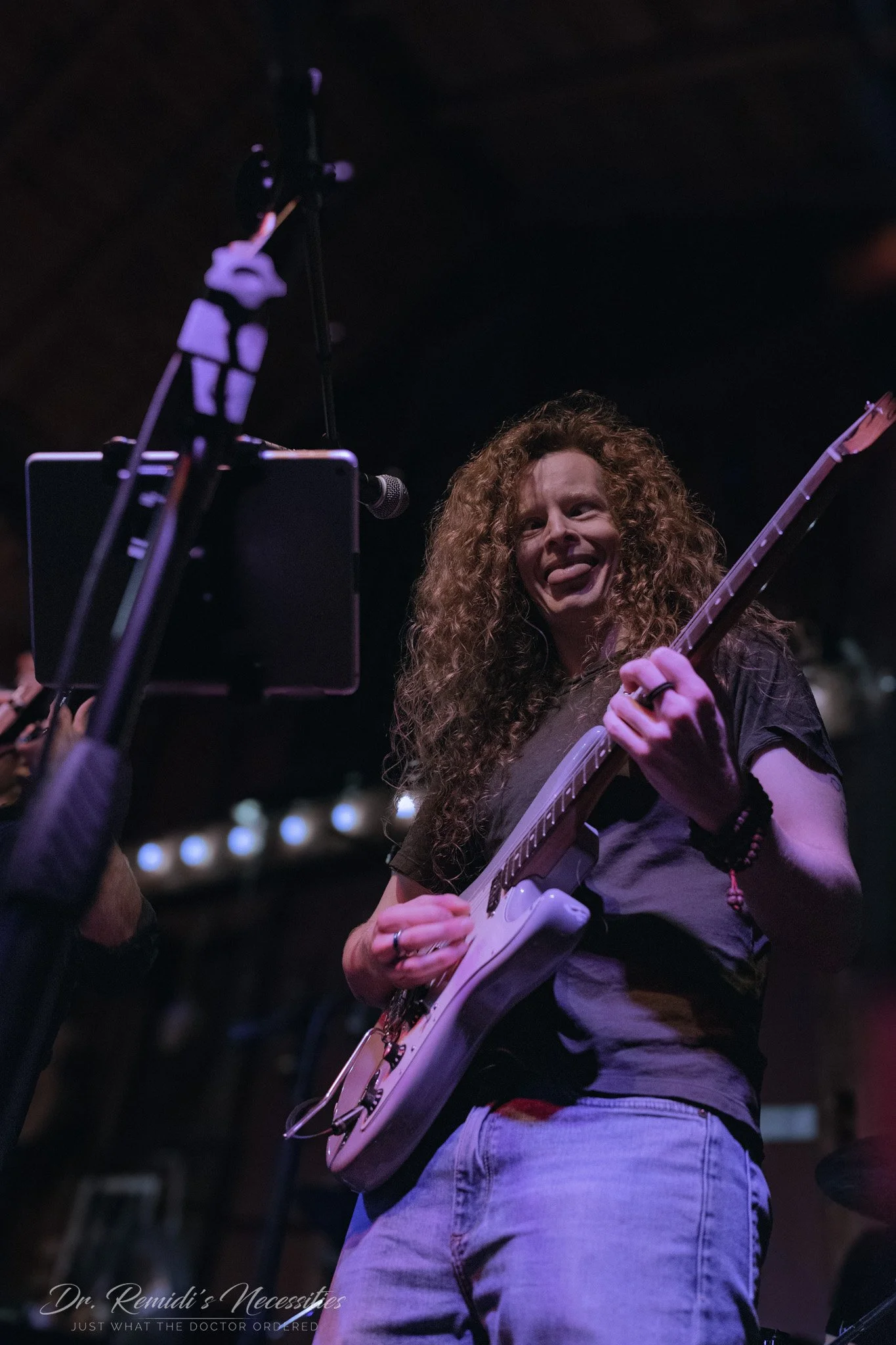A woman with curly hair playing an electric guitar on stage with a dark background and stage lights.