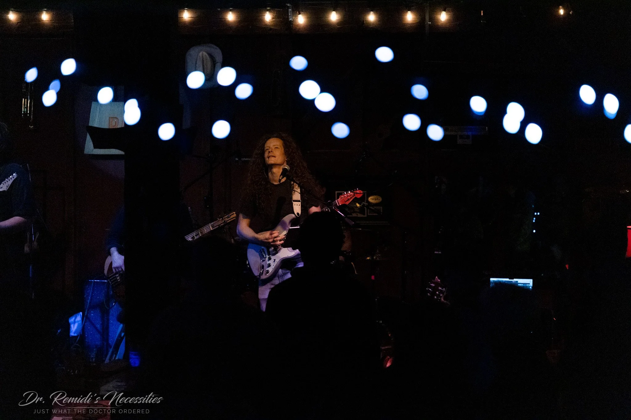 A female guitarist with curly hair performing on stage in a dimly lit venue decorated with string lights and illuminated orbs hanging from the ceiling, with silhouettes of audience members in the foreground.