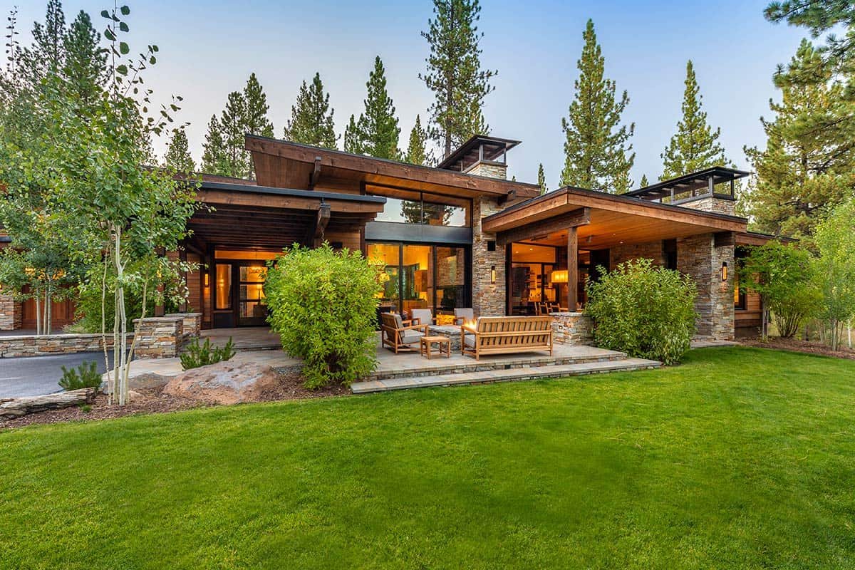 Modern house with stone and wood exterior, surrounded by trees, featuring a patio with outdoor furniture, set on a lush green lawn.