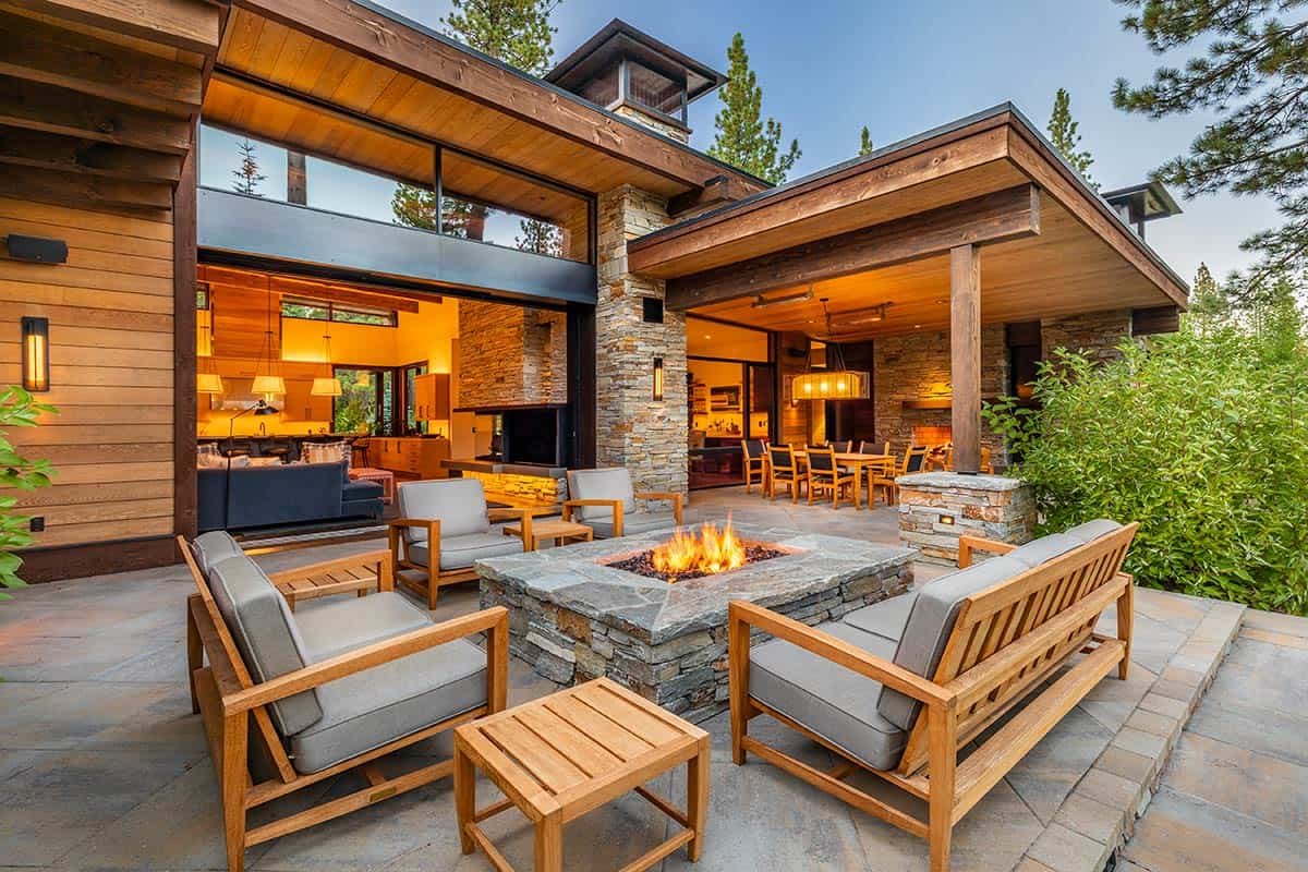 Modern patio with wooden furniture and a fire pit, open view to interior dining and living areas with large windows, surrounded by trees.