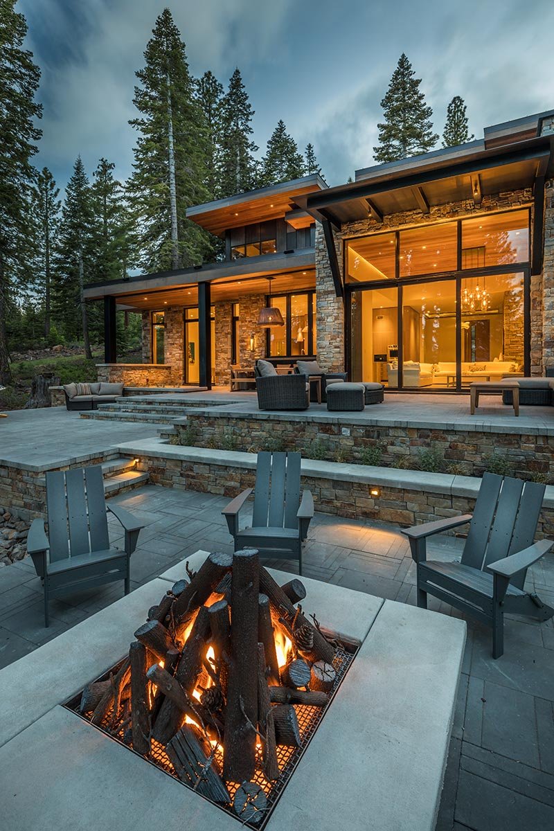 Modern house with large windows and outdoor patio with fire pit, surrounded by tall pine trees.