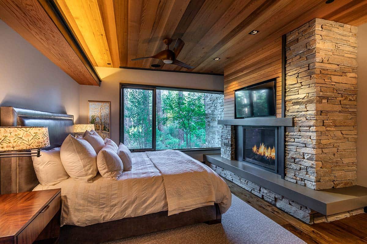 Modern bedroom with wooden ceiling, large windows, bed with white bedding, stone fireplace, and mounted TV.