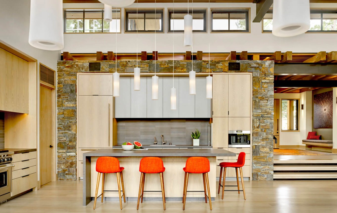 Modern kitchen with wooden cabinets, stone accents, red barstools, hanging pendant lights, and a large island.