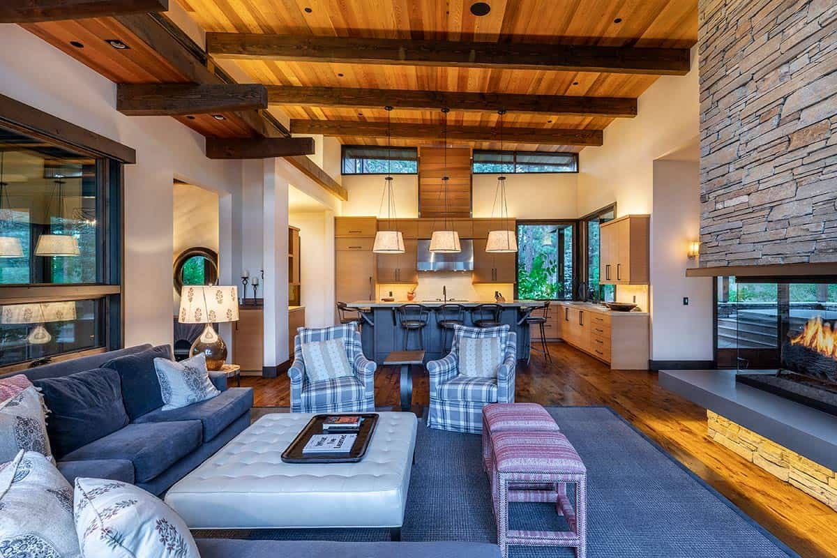 Modern open-concept living space with high wooden ceilings, featuring a cozy seating area with a gray sofa, plaid armchairs, and a tufted ottoman. A stone fireplace is visible to the right, and a spacious kitchen with an island, bar stools, and penda