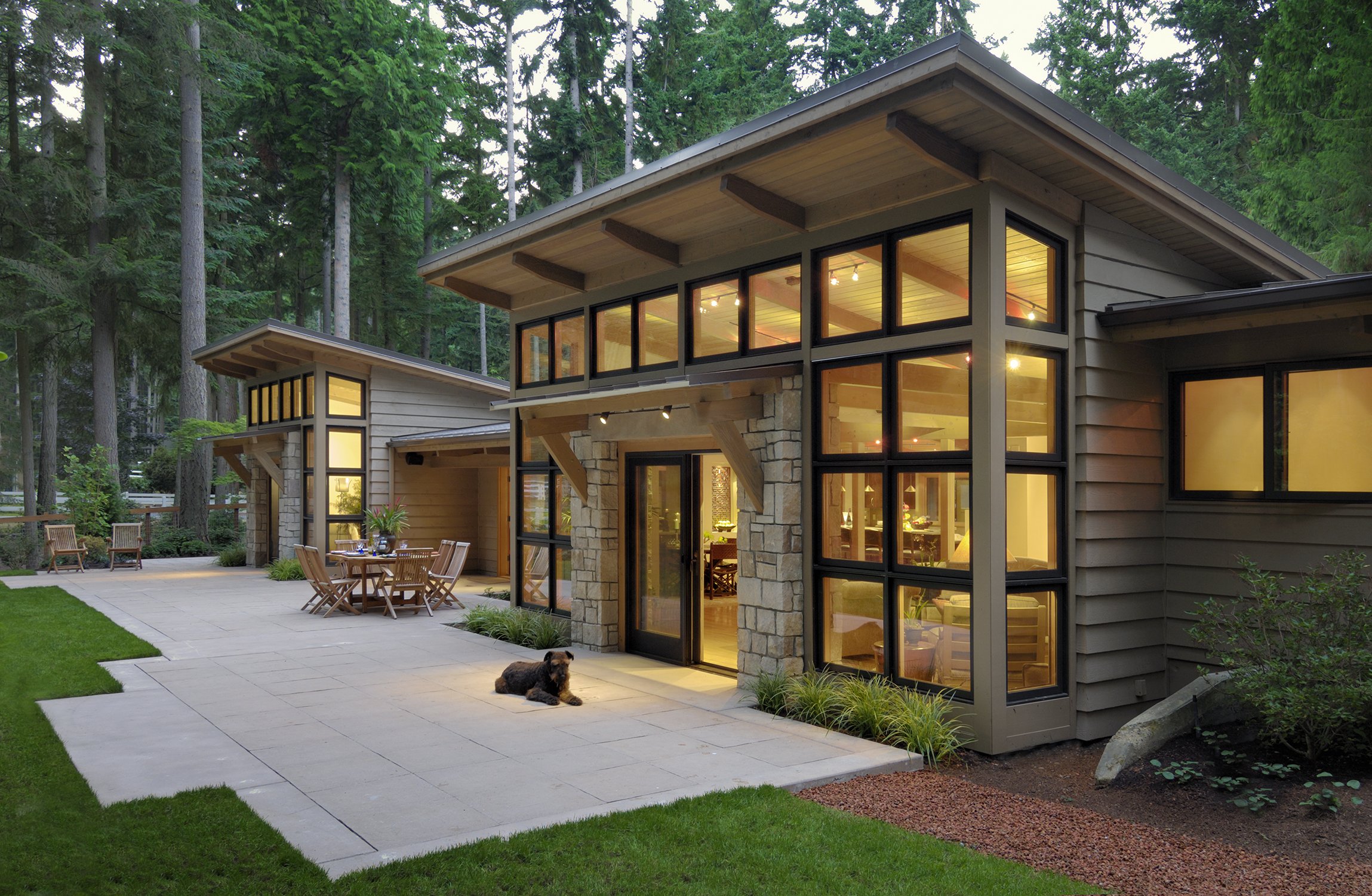Modern house with large windows, patio area, outdoor seating, and trees in the background.