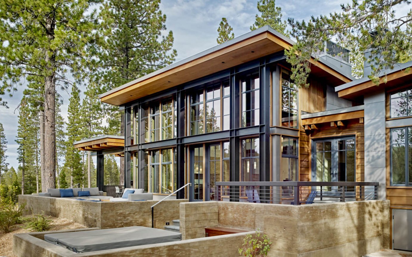 Modern house exterior with large glass windows, wood accents, outdoor seating, surrounded by pine trees.