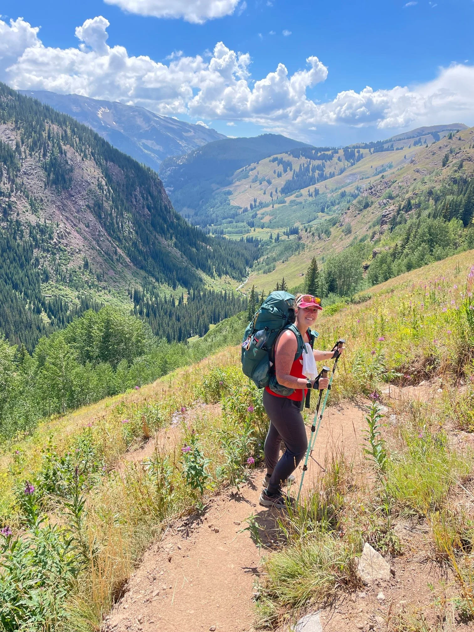 A woman hiking on a mountain trail with a backpack and trekking poles, surrounded by green vegetation, with a backdrop of mountains, trees, and a partly cloudy sky.