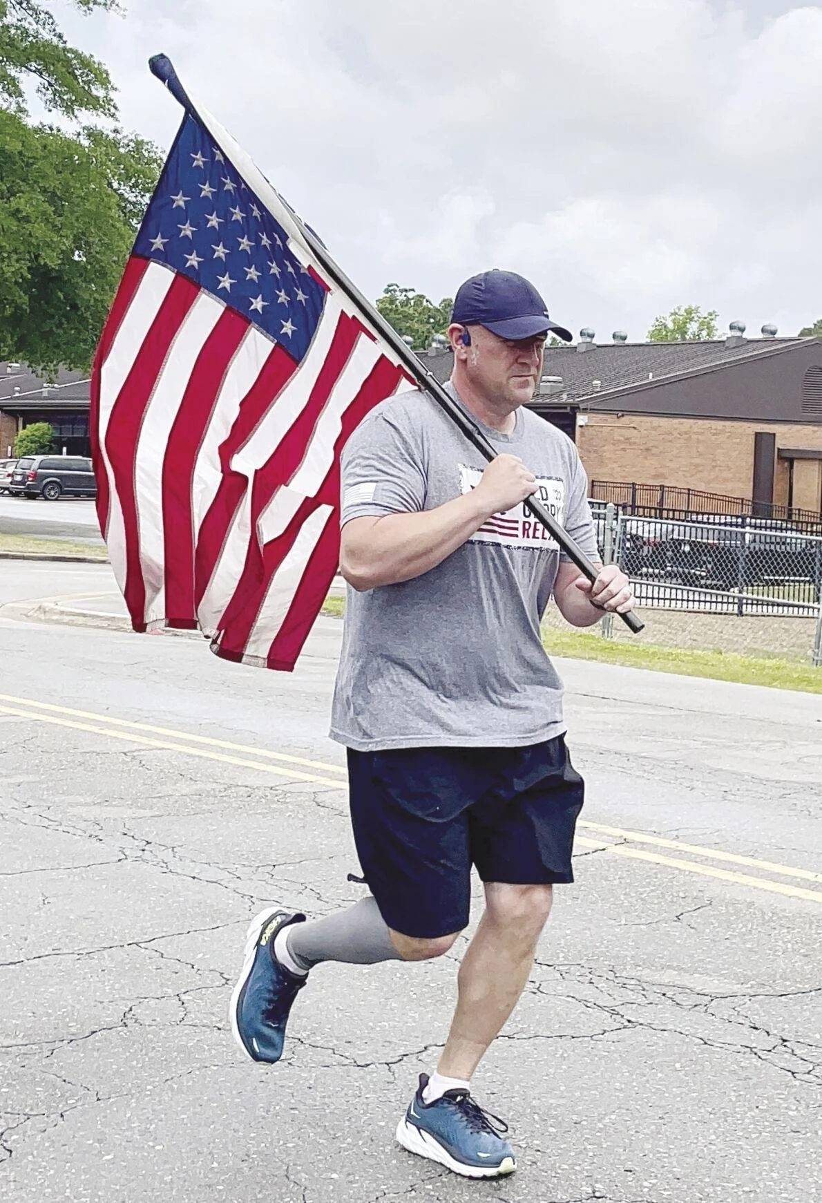A man running on a street while carrying an American flag, wearing a gray t-shirt, black shorts, a navy cap, and running shoes.