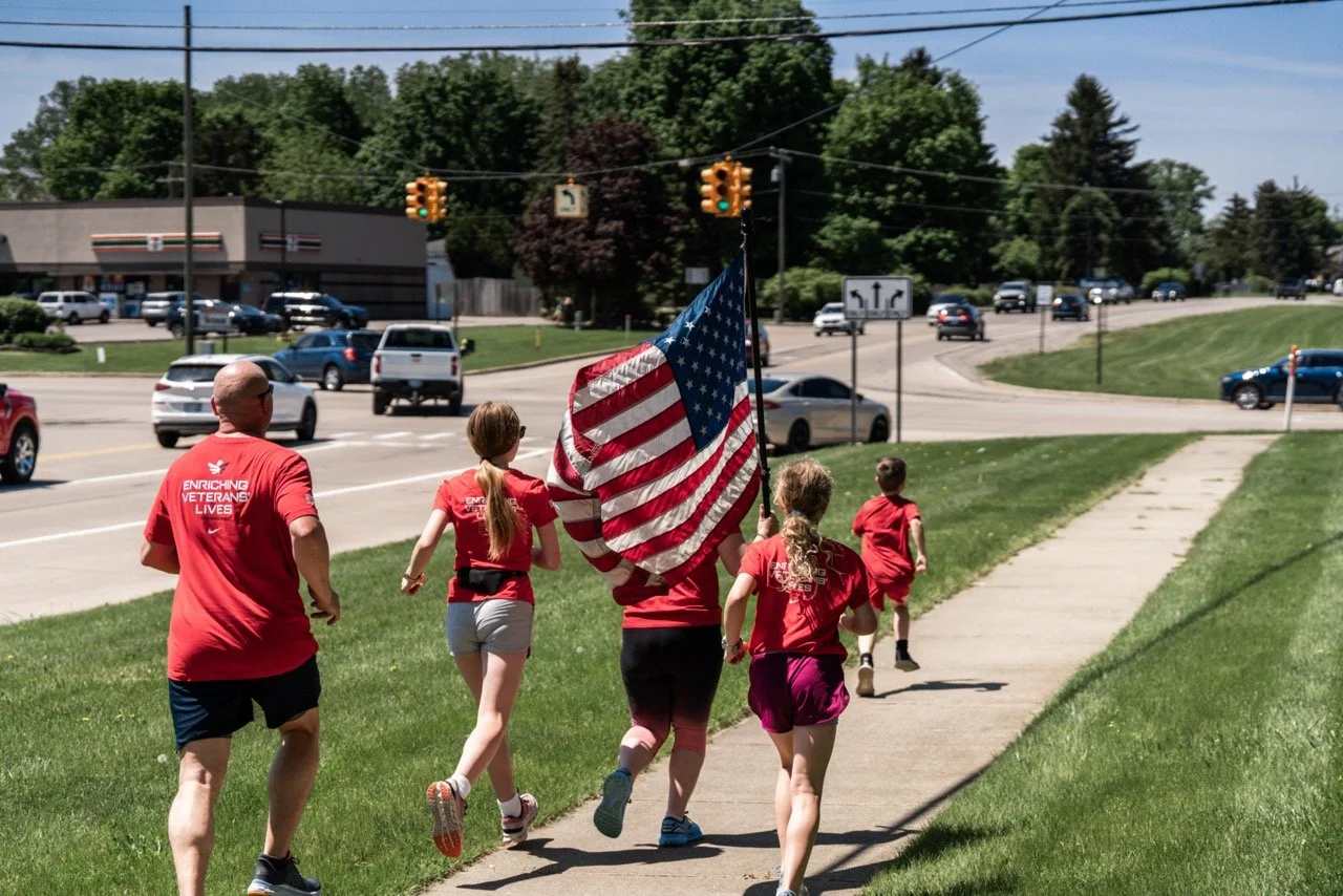 Group of people running on a sidewalk, holding an American flag. They are wearing red T-shirts, some with the words "Enriching Veterans Lives," on a sunny day with trees and traffic at an intersection in the background.