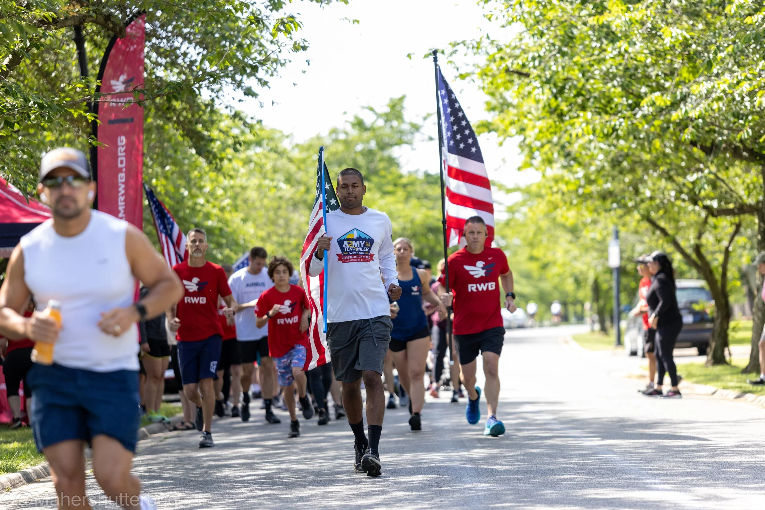 People participating in a patriotic run holding American flags on a tree-lined street.
