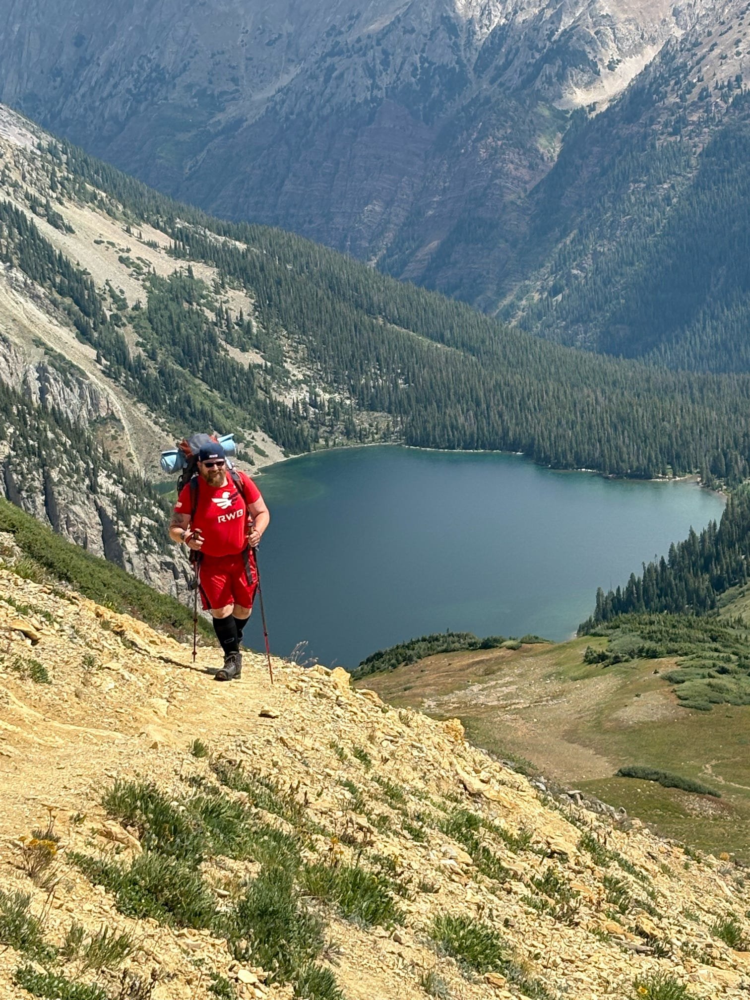 A hiker with a backpack trekking on a mountain trail near a lake, surrounded by green and rocky terrain with a mountain range in the background.