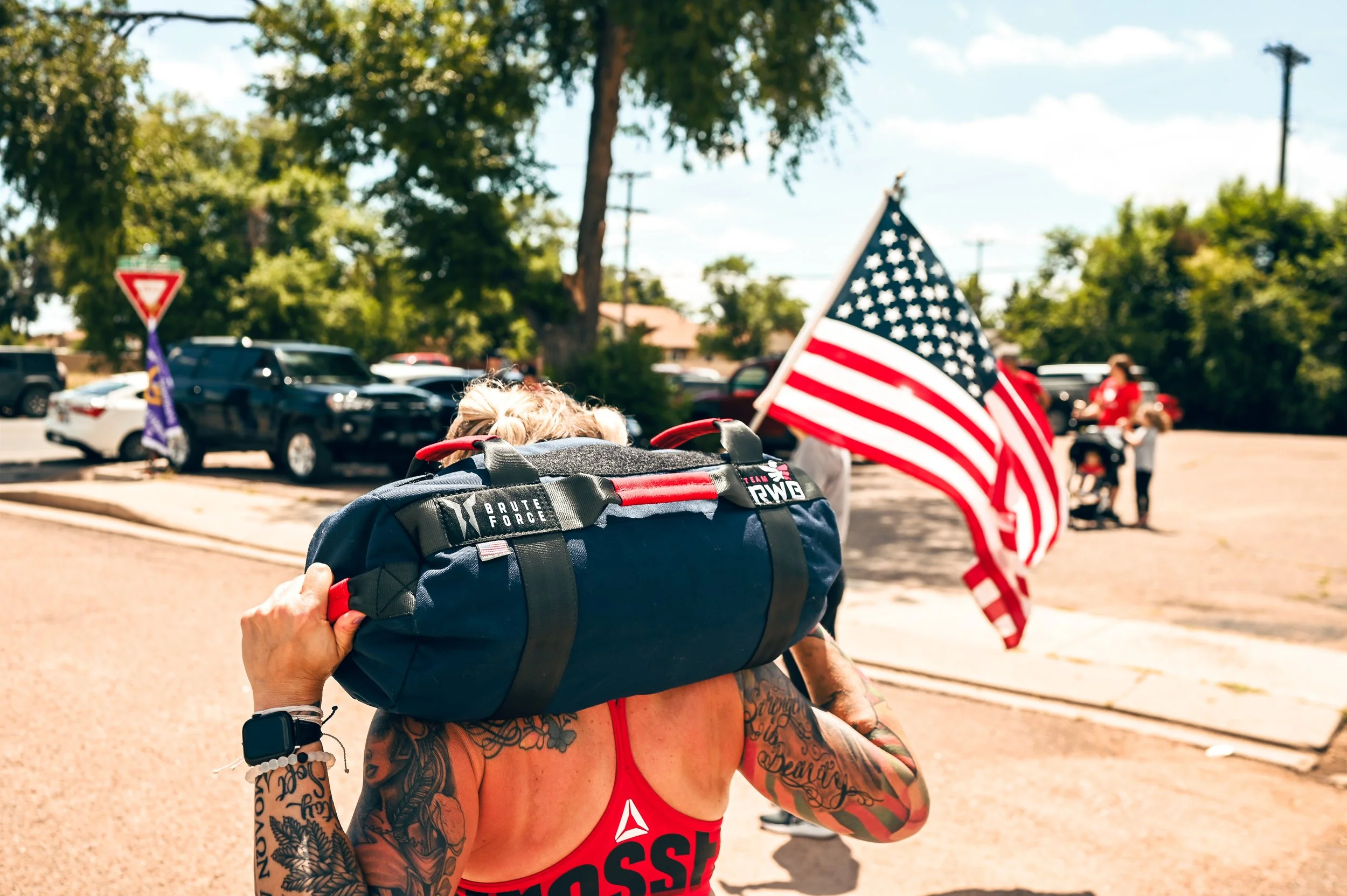 A woman wearing a red tank top with tattoos on her arms carries a blue duffel bag on her shoulder at an outdoor event, with American flags and people in the background.