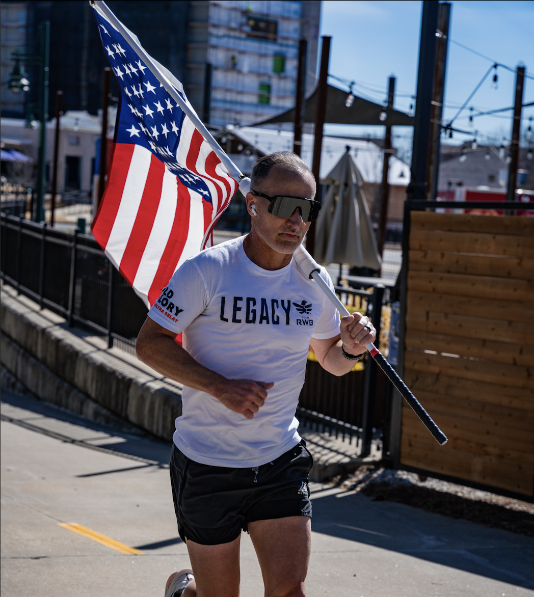 Man running outdoors during daytime, carrying an American flag and wearing sunglasses and a white T-shirt that reads 'LEGACY'.