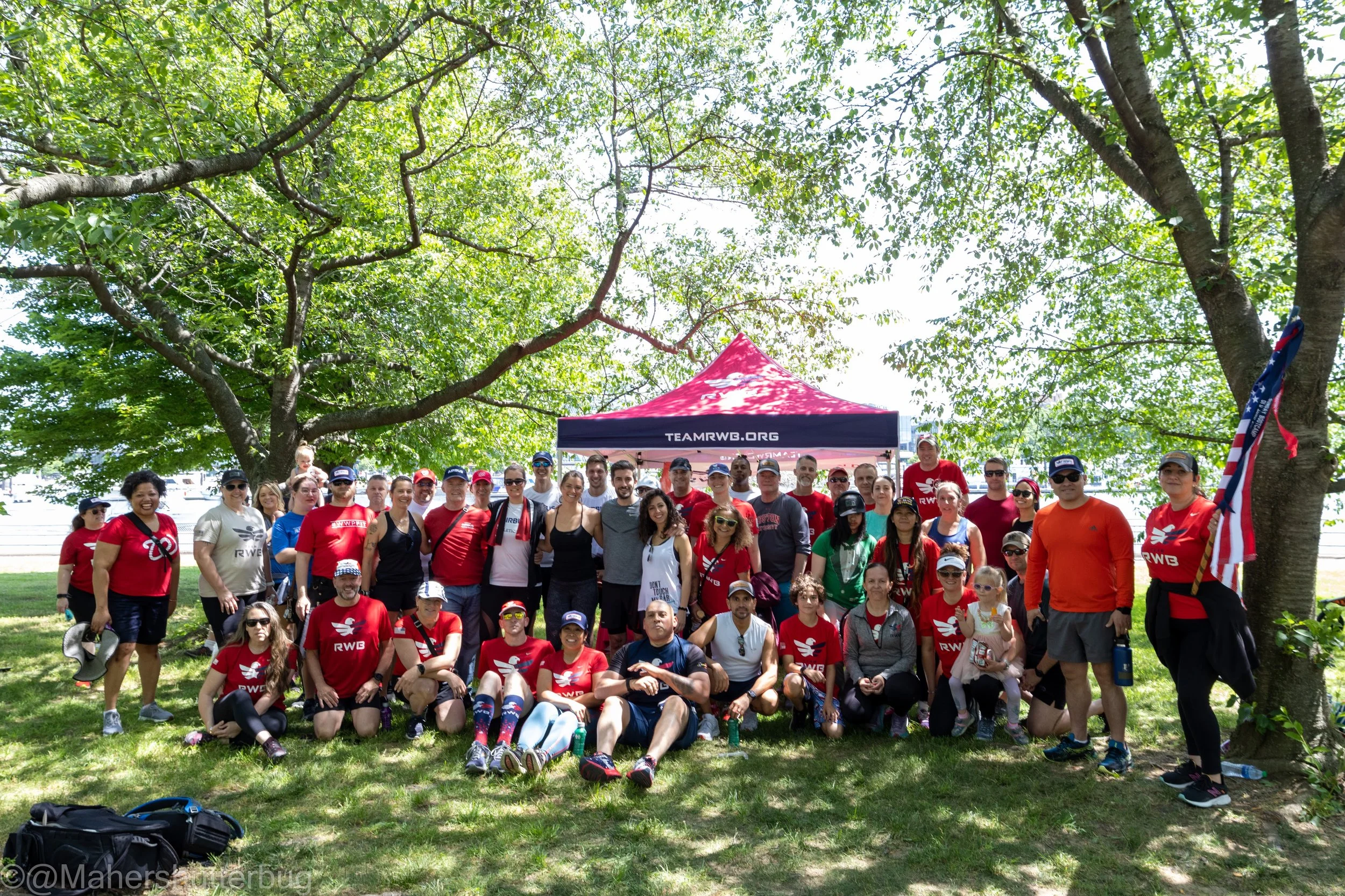 A large group of people gathered outdoors under trees, posing for a photo in front of a red tent with