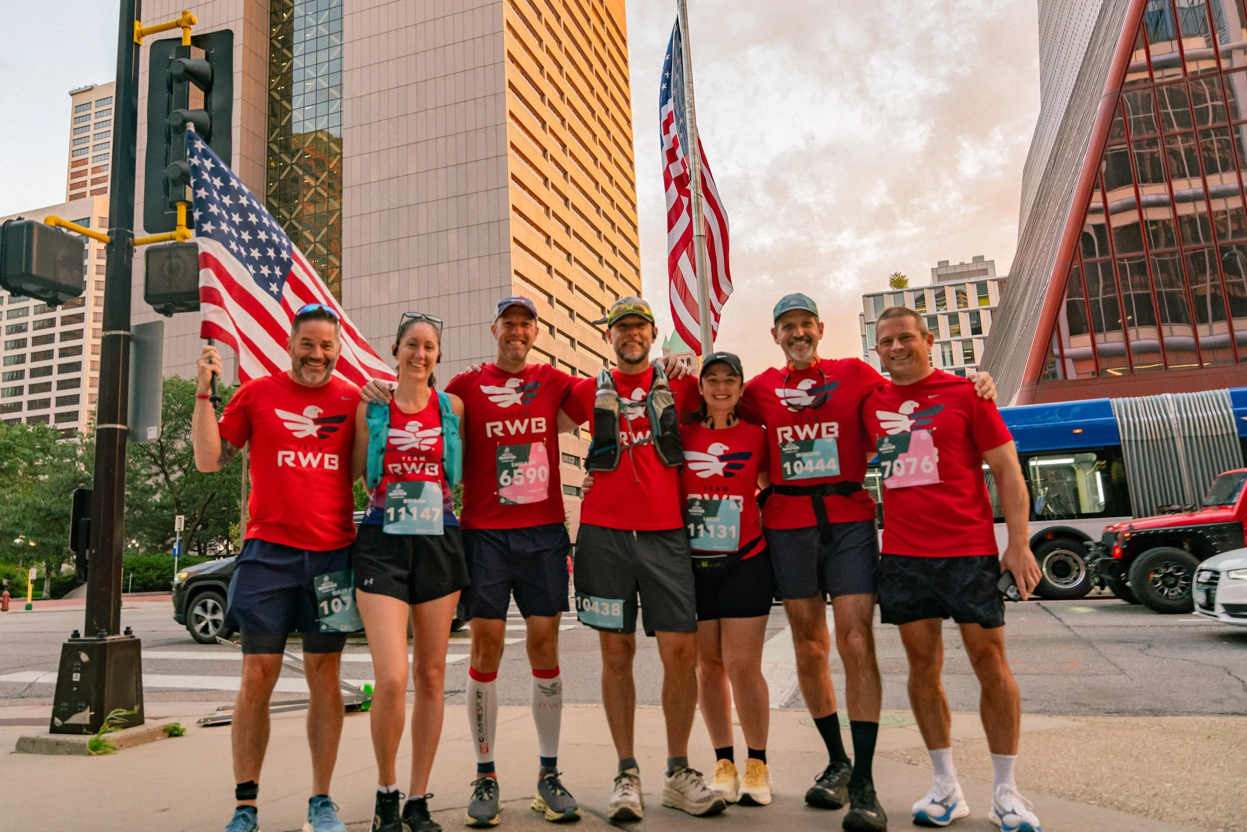 Seven runners in red shirts and shorts standing together on city street with tall buildings and American flags, celebrating after a race.
