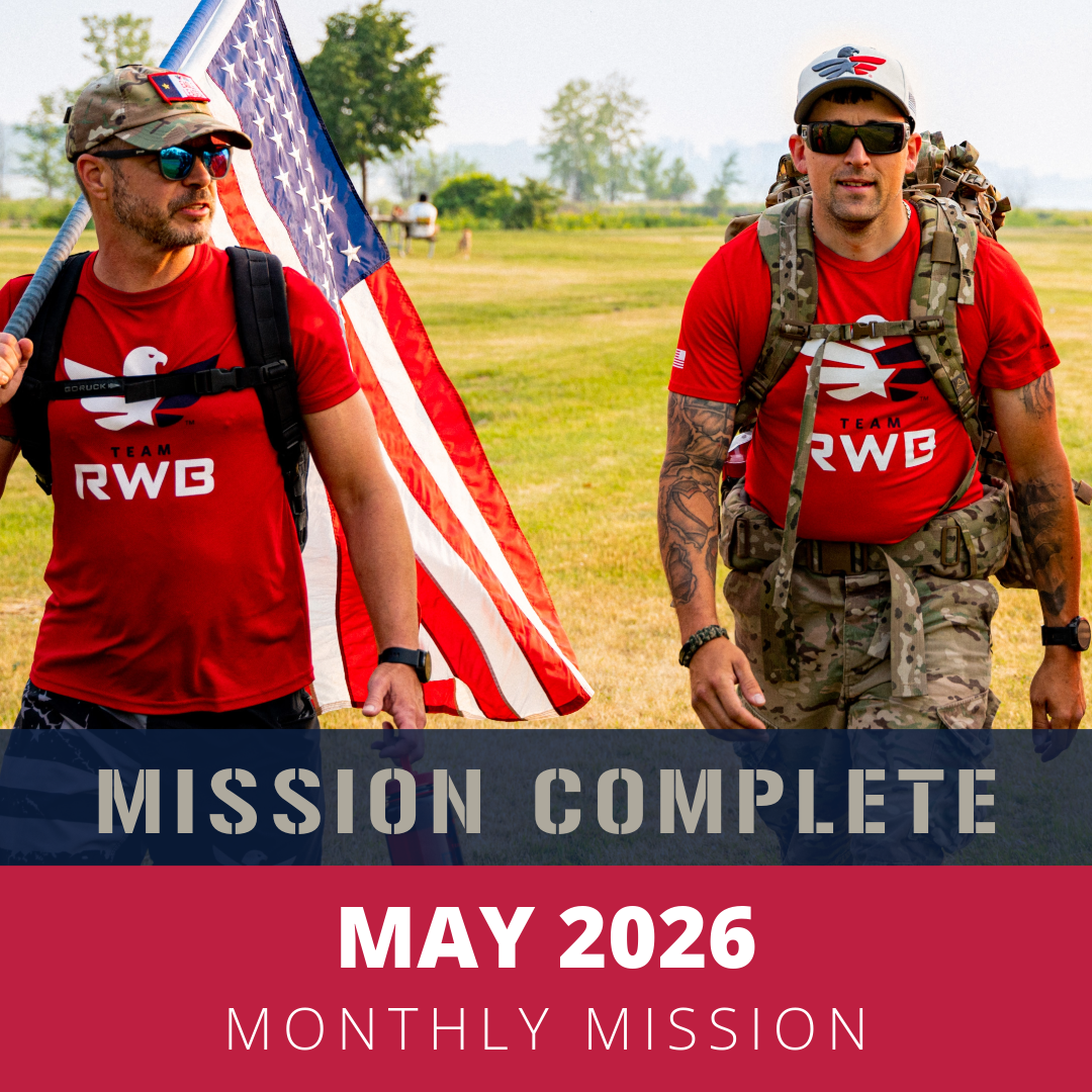 Two men in red shirts with 'Team RWB' logo, wearing backpacks and sunglasses, walking across a grassy field with an American flag. One holds a water bottle, both appear to be participating in a May 2026 mission or event.