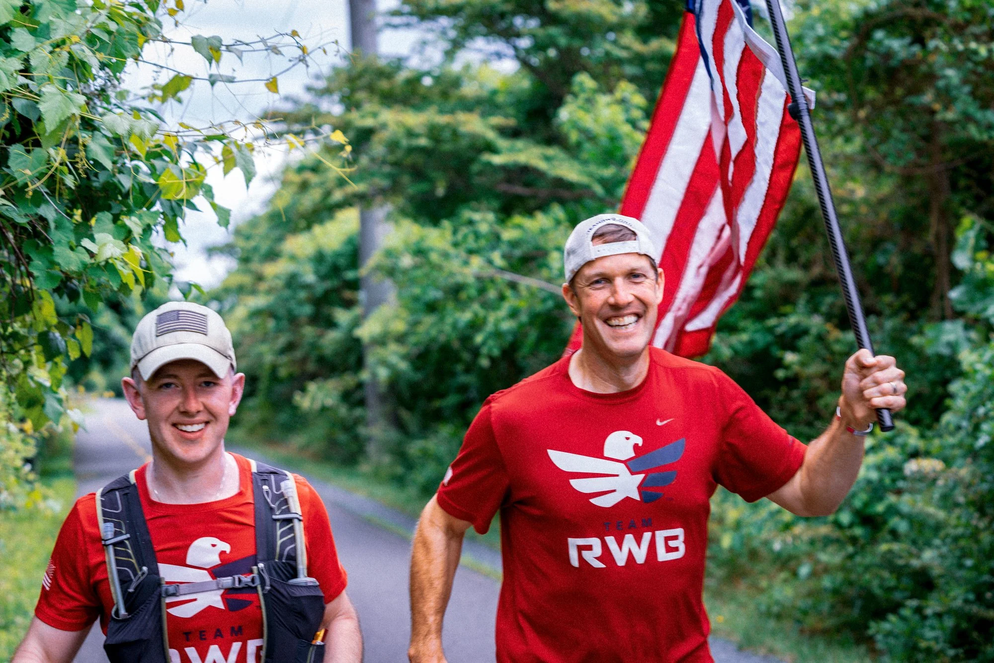 Two men running outdoors on a trail surrounded by green trees, both wearing red shirts with a white eagle logo and the words 'TEAM RWB'. The man on the right is smiling broadly and holding an American flag, while the man on the left is smiling and wearing a backpack.