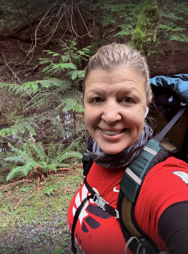 A smiling woman with her hair in a ponytail hiking in the PNW wearing a red t-shirt that says Team RWB