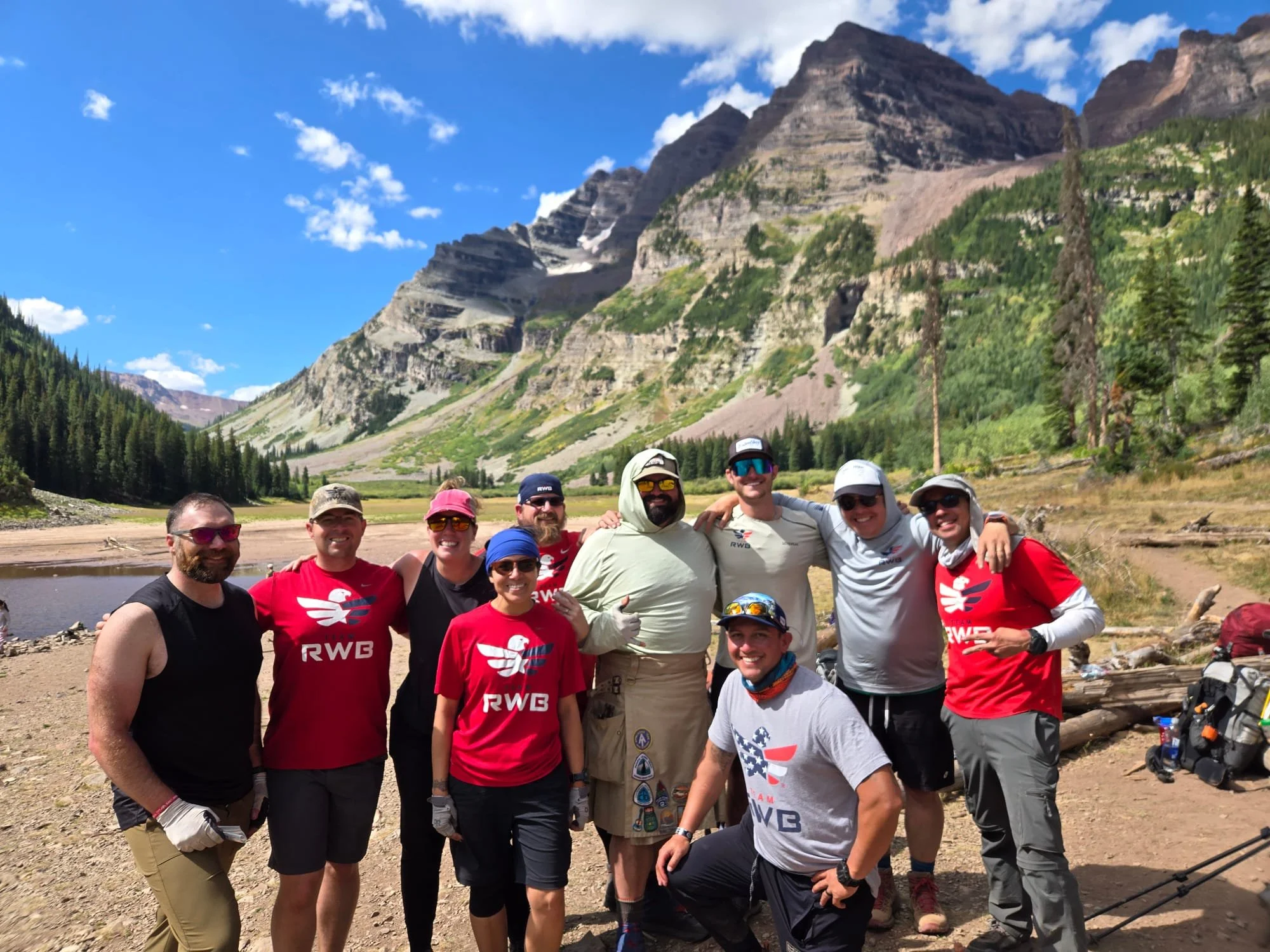 A group of people posing outdoors in a mountainous area with tall peaks covered in greenery and patches of snow in the background. The group appears to be hikers or adventurers, dressed in casual outdoor clothing and smiling at the camera.