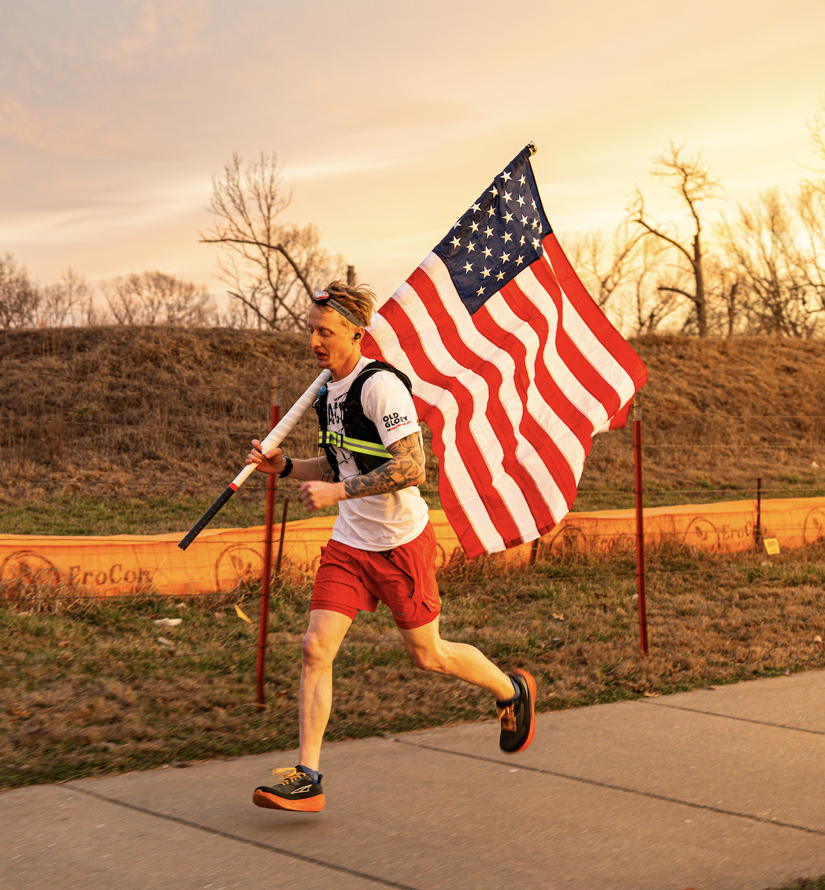 A man running outdoors at sunset, holding a large American flag on a pole, dressed in athletic gear with a white shirt, red shorts, and running shoes.