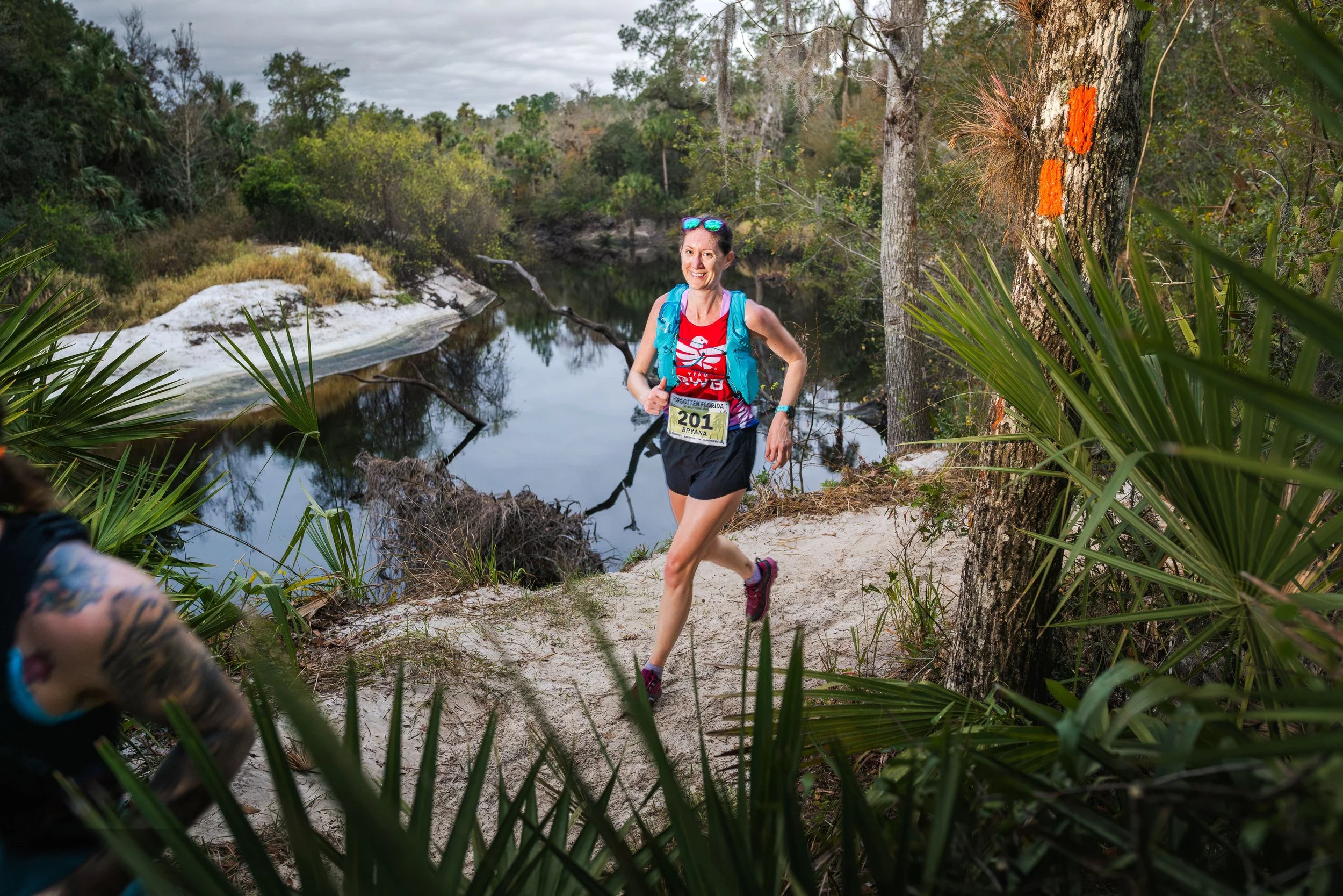A female runner smiling while running in a wooded outdoor setting.