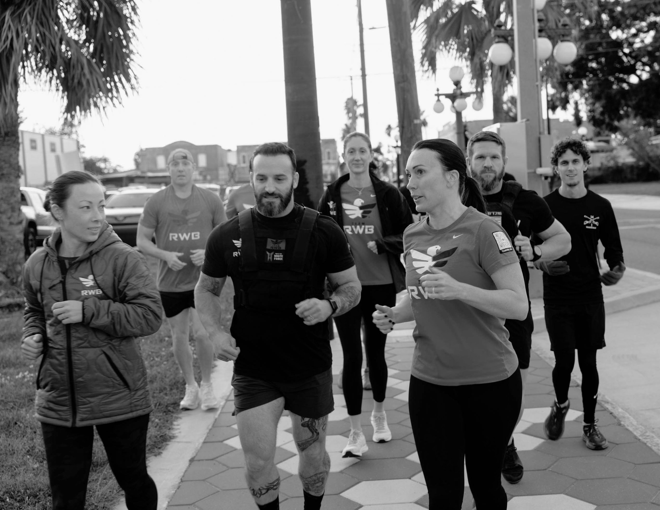 Group of seven runners, four women and three men, jogging along a sidewalk outdoors on a cloudy day, dressed in athletic clothing, with trees and streetlights in the background.