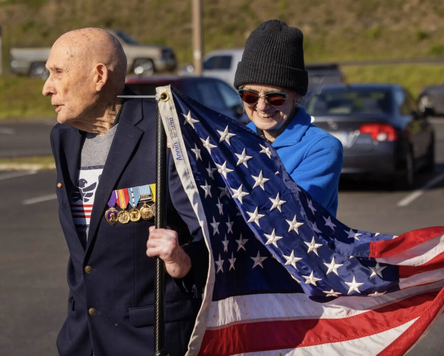 An elderly man with medals on his blazer is being assisted by a woman in a blue hoodie and black beanie as they hold an American flag.