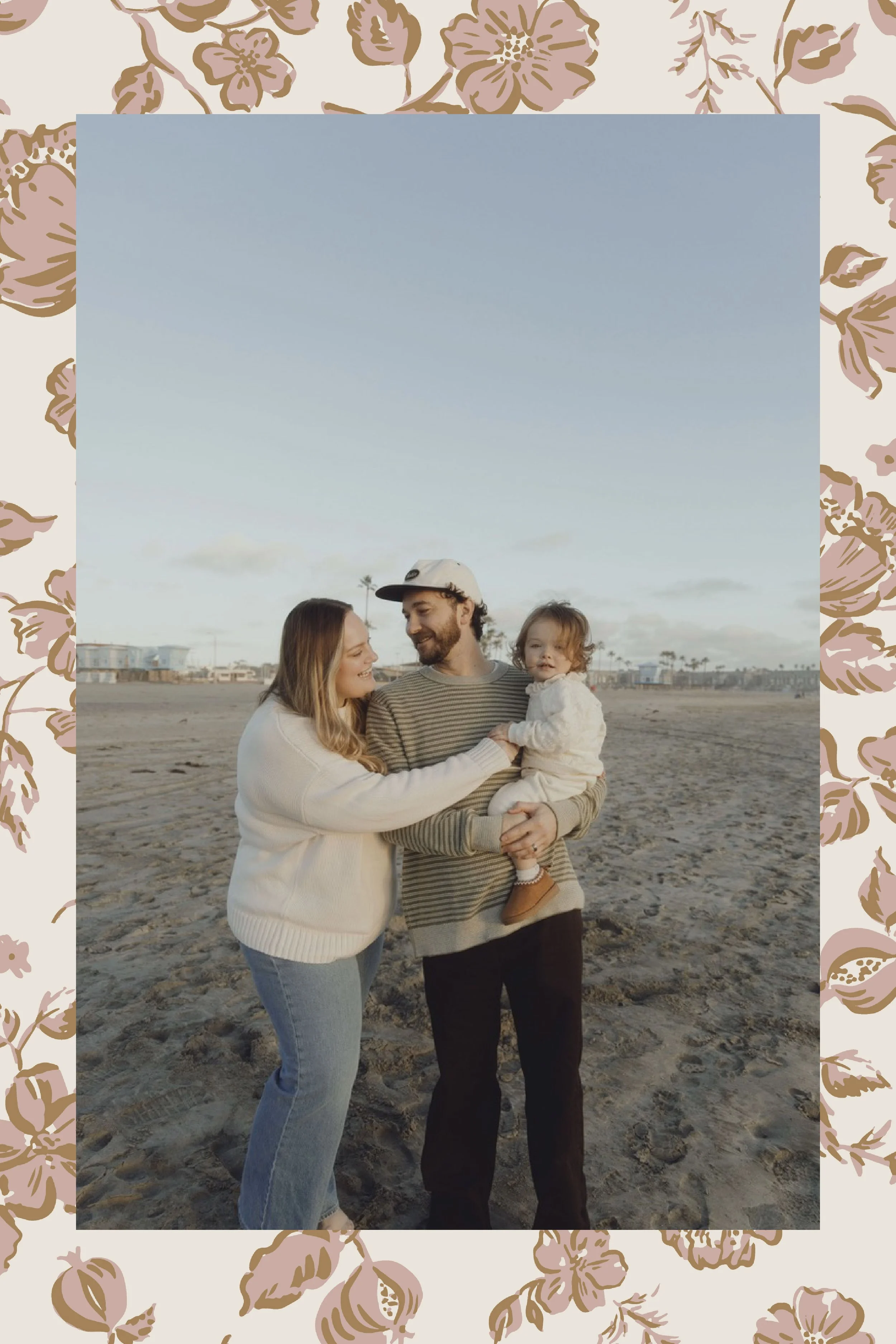 A family of three, including a woman, a man, and a young child, standing together on a beach during sunset, smiling and looking at each other.