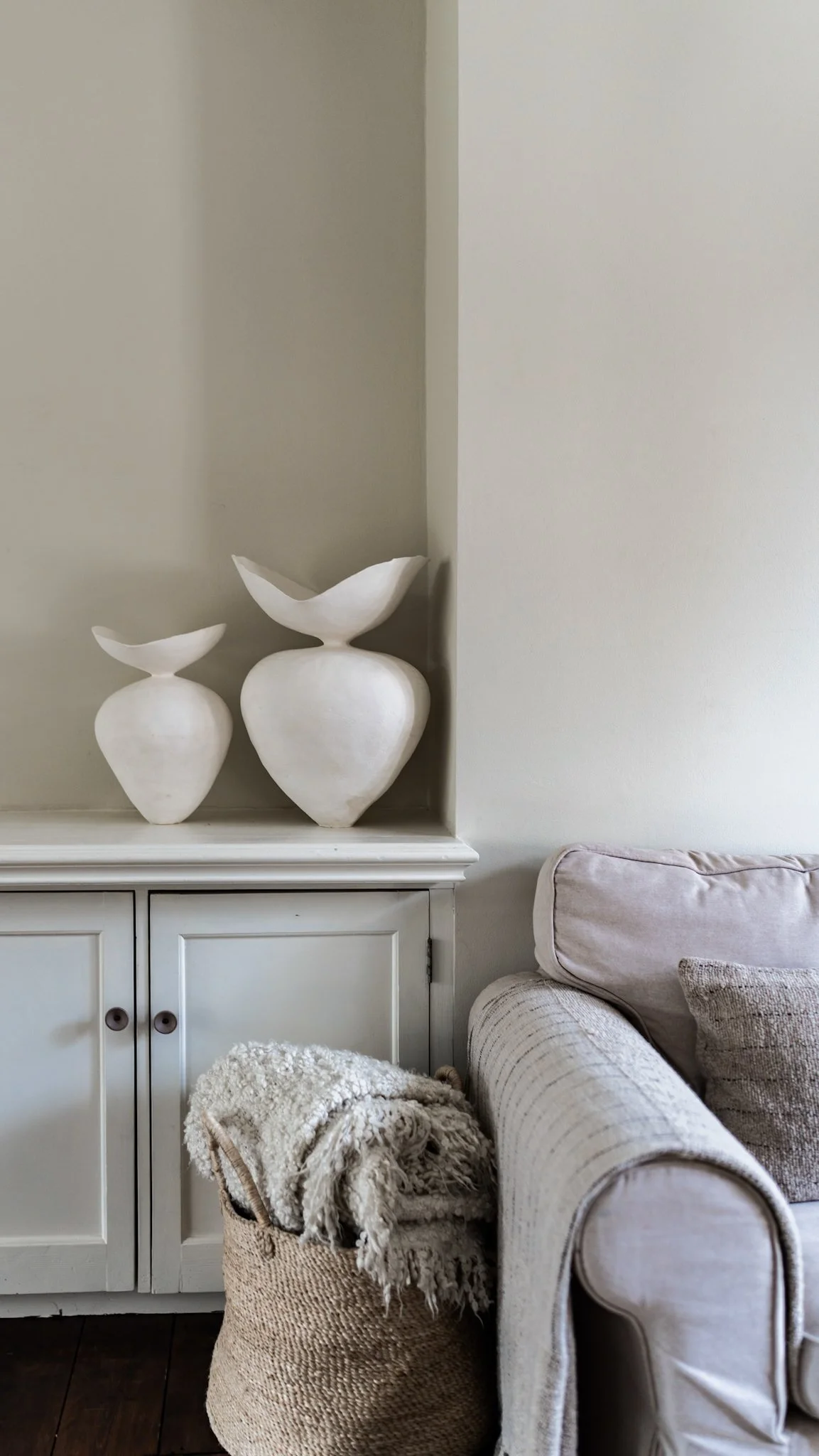 Decorative white vases on a white cabinet placed in a corner near a beige sofa.