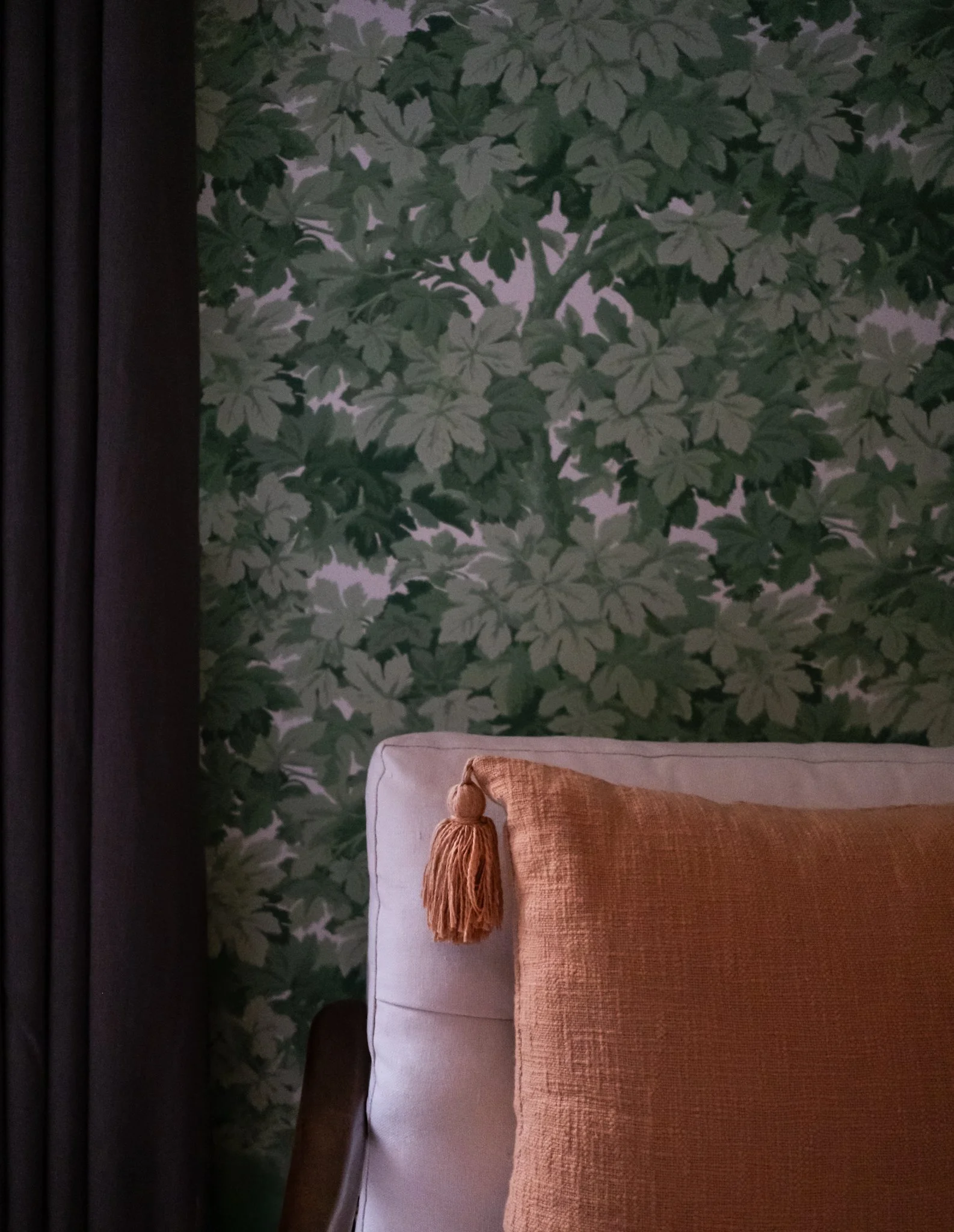 A beige upholstered headboard with an orange cushion and a tassel, placed against a leafy green wallpapered wall with dark curtains on the side.