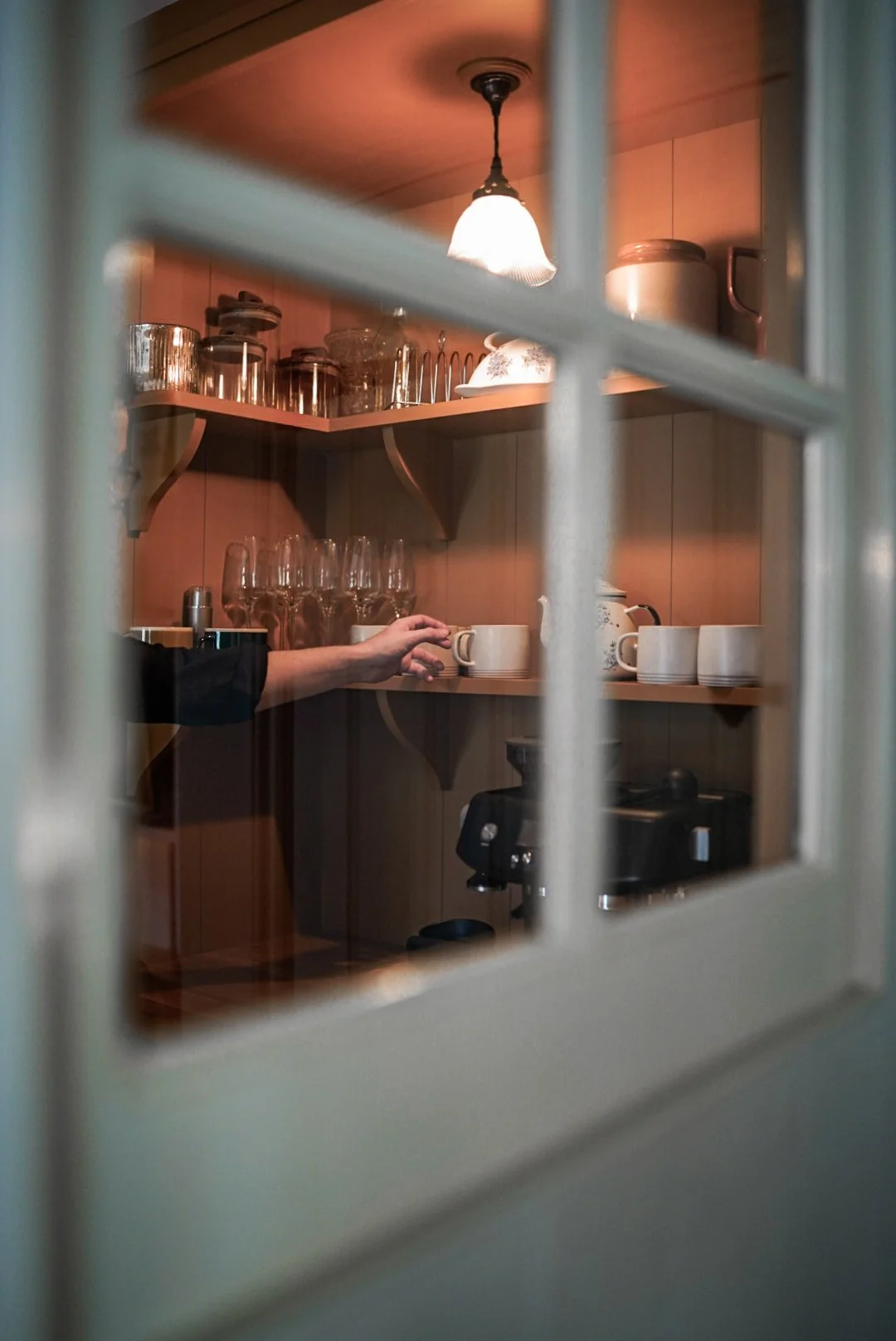 Kitchen cabinet viewed through a window, with glasses, mugs, and a coffee machine inside.