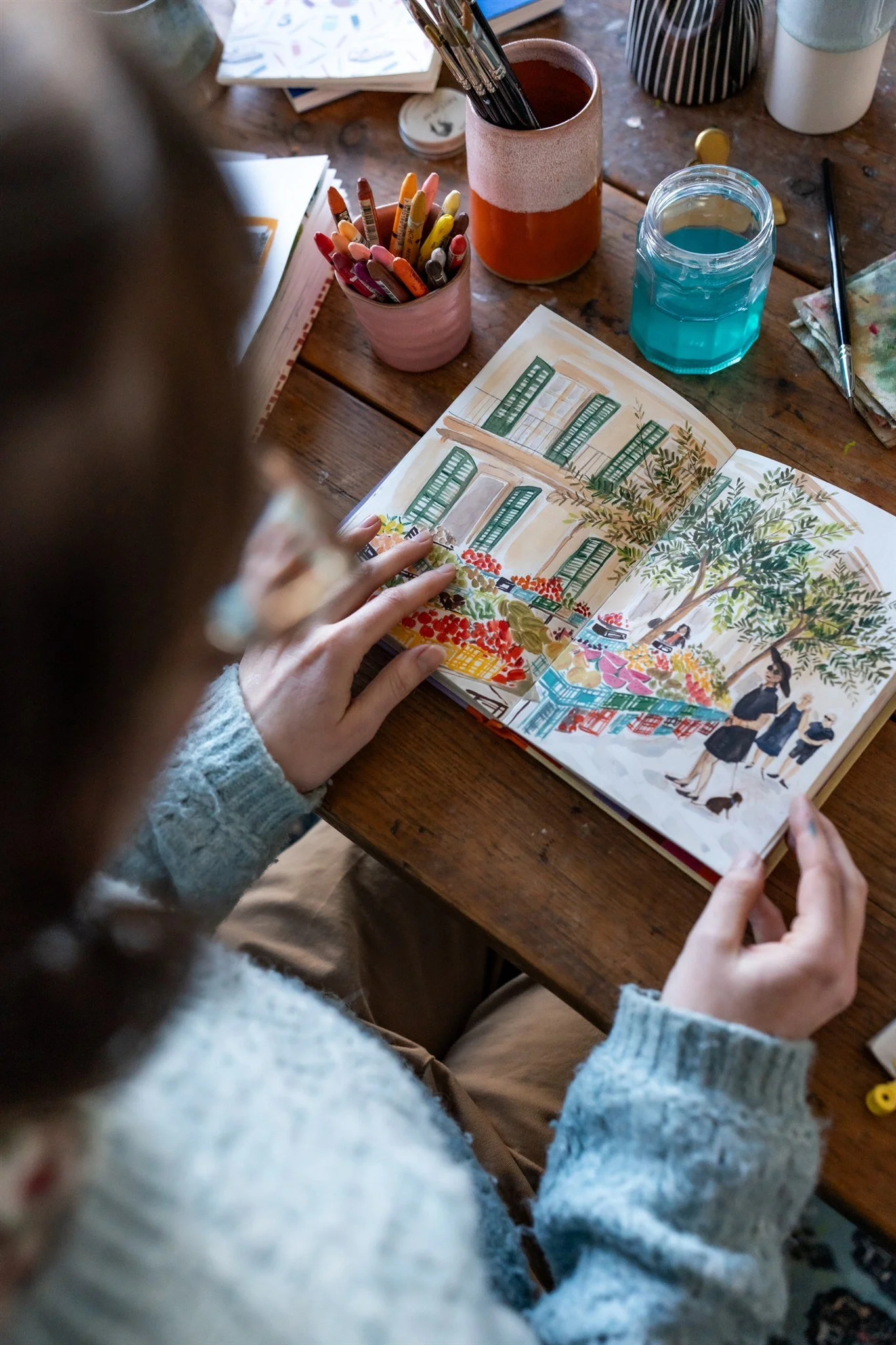 Person reading a colorful illustrated book at a wooden table with art supplies, including markers, water glasses, and a paintbrush.