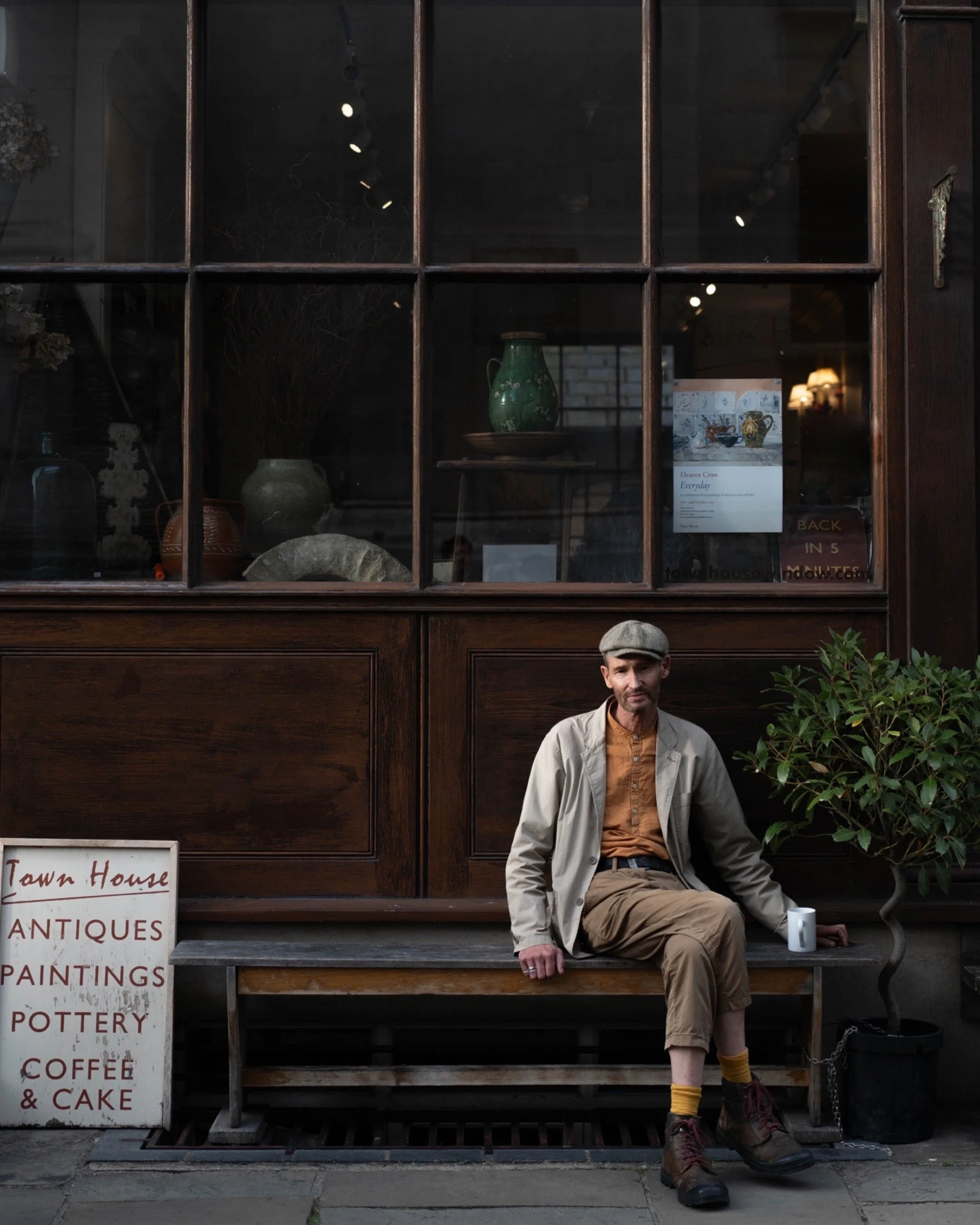 A man sitting on a wooden bench outside a vintage shop with a sign advertising antiques, paintings, pottery, coffee, and cake. The shop has a large glass window showcasing various pottery and decorative items. The man is wearing a beige coat, rust-colored shirt, khaki pants, and a gray cap, with a white mug placed on the bench next to him. There is a small potted tree beside him.
