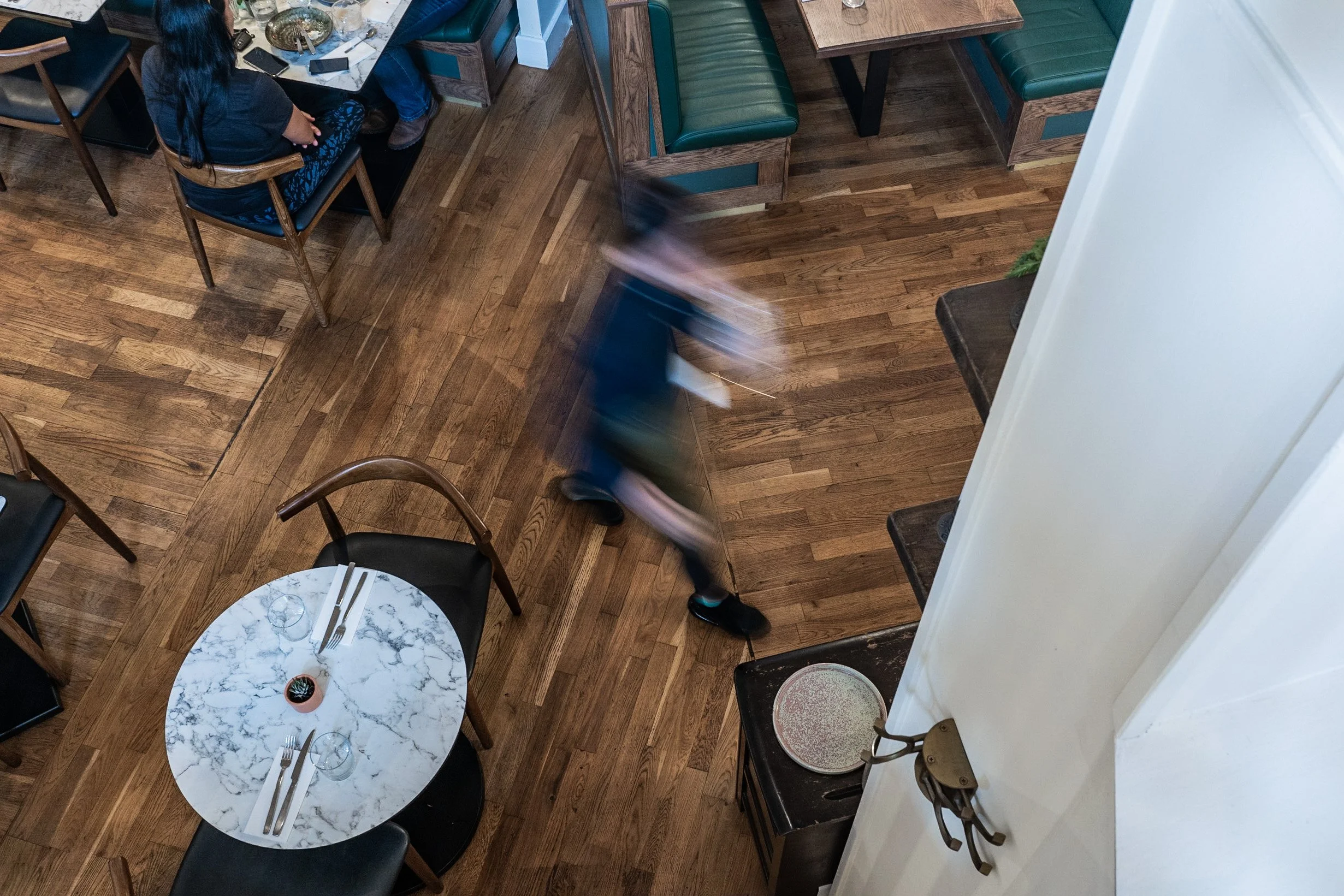 A bird's eye view of a restaurant interior with wooden flooring, tables and chairs, including a marble round table set with utensils and glasses, a woman sitting at another table, and a person walking past.