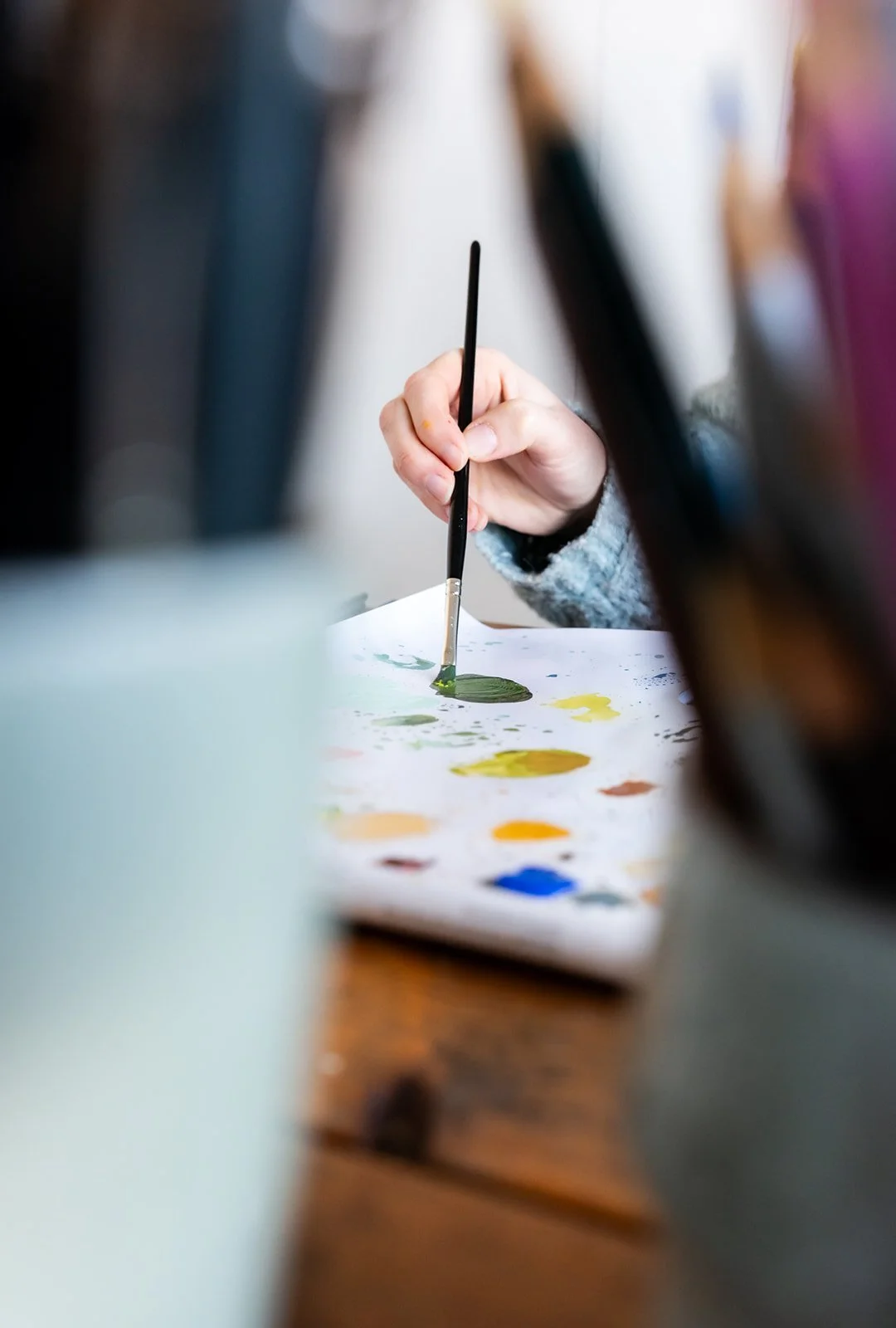 A person using a small paintbrush to apply green paint on paper, with colorful paint spots on the table.