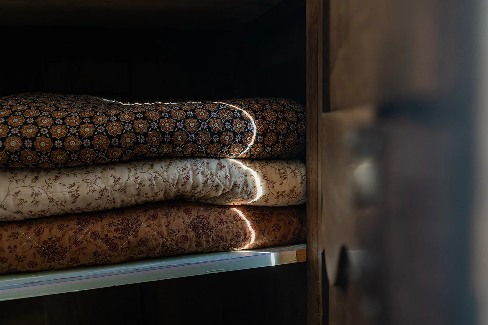 Stacked mattresses with patterned covers stored on a shelf with sunlight peeking through.