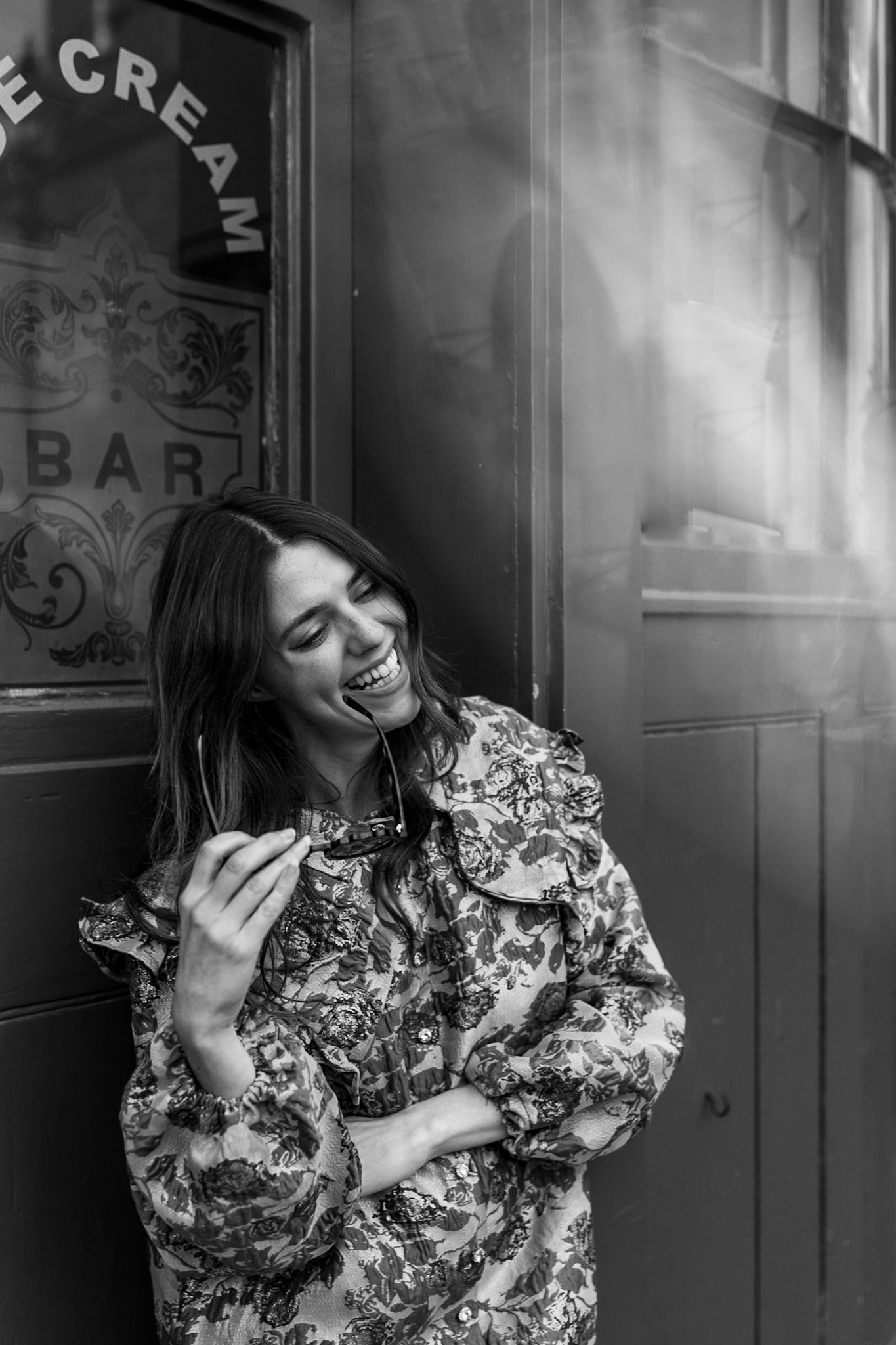 A woman with long hair, laughing happily, holding sunglasses near her face, standing against a dark wall with a window that has 