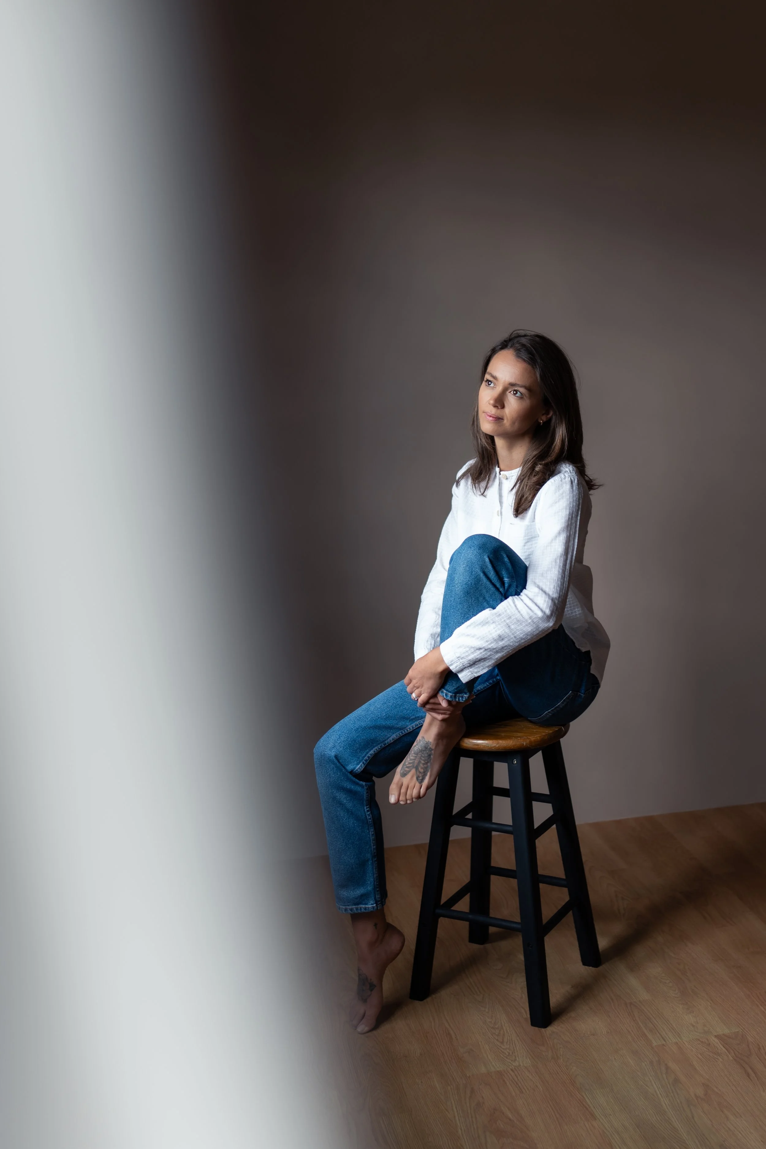 A woman with long brown hair sitting barefoot on a wooden stool, wearing a white shirt and blue jeans, in a studio with a light-colored wall and wooden floor, gazing thoughtfully to her left.
