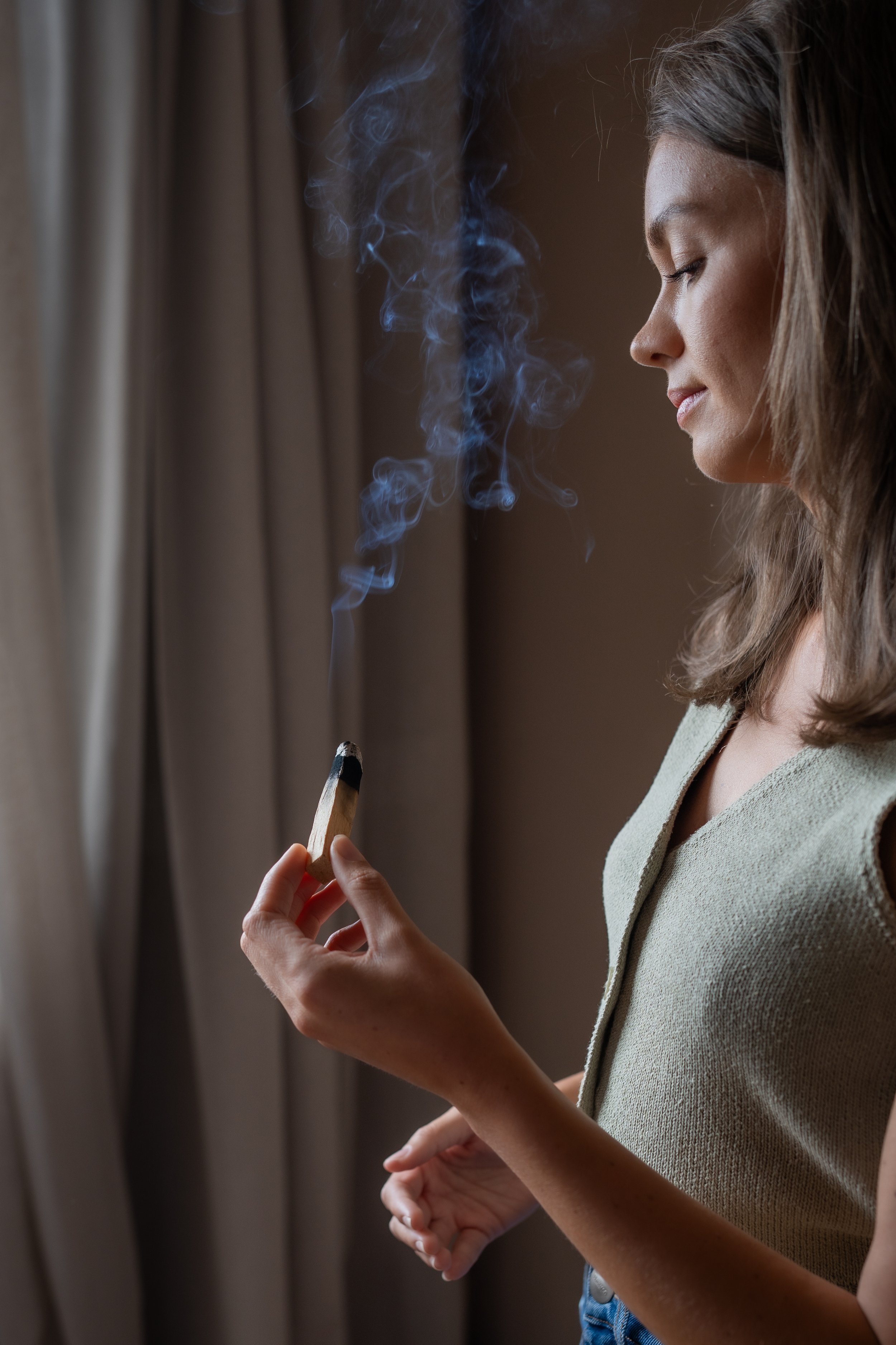 A woman with shoulder-length grey hair is holding a burning stick of sage close to her face, with smoke rising from it.