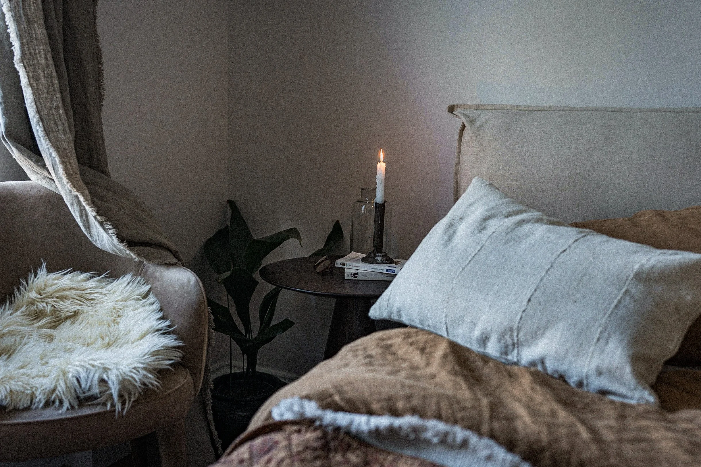 A cozy living room corner with a beige armchair, a fluffy white throw, a bed with large beige and brown pillows, a small round black side table with a lit candle, a glass jar, and a plant in the background.