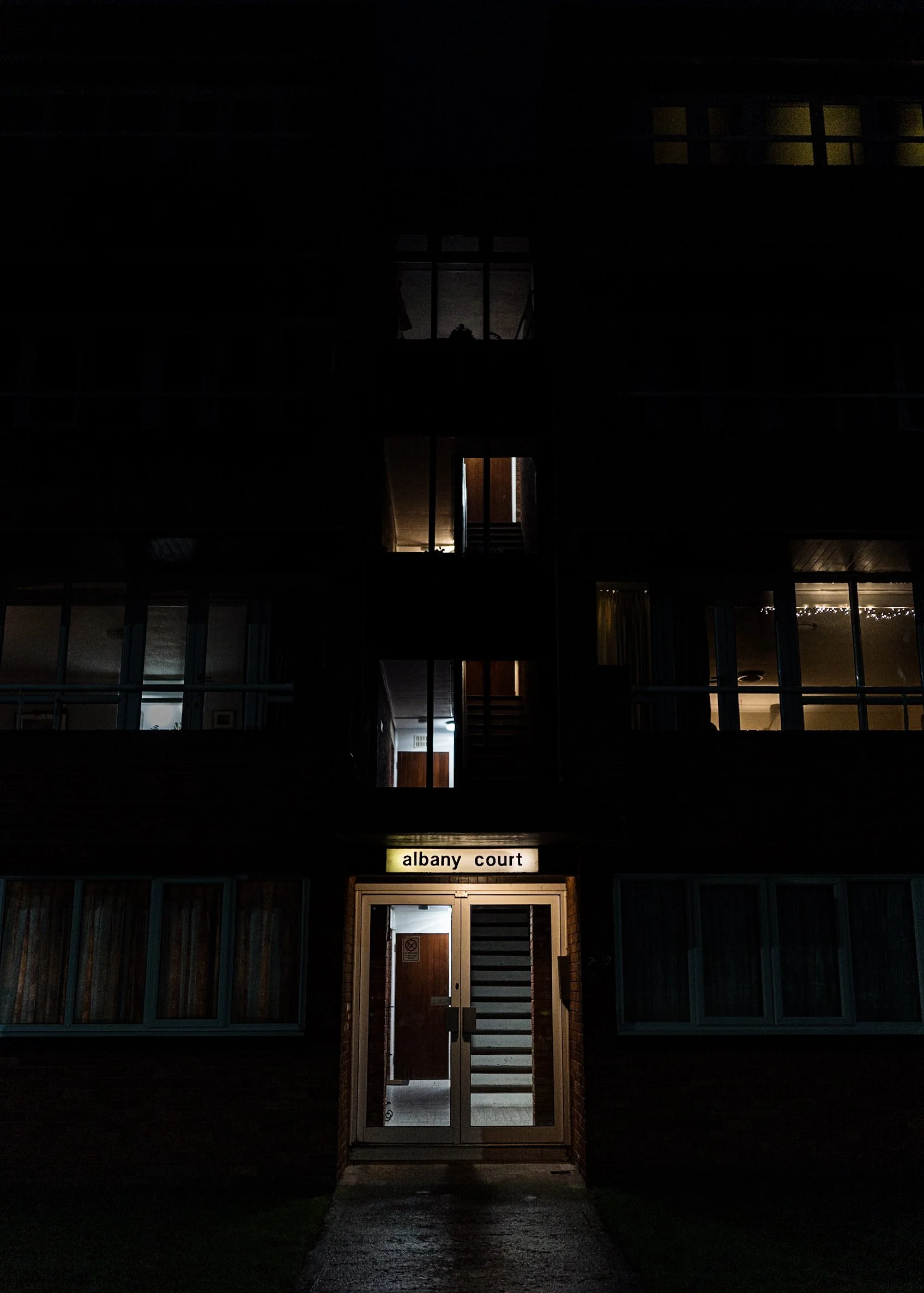Nighttime photograph of a multi-story apartment building with illuminated sign reading 'albany court' above the entrance door. The building has several lit windows, and external stairs are visible through the open entrance.