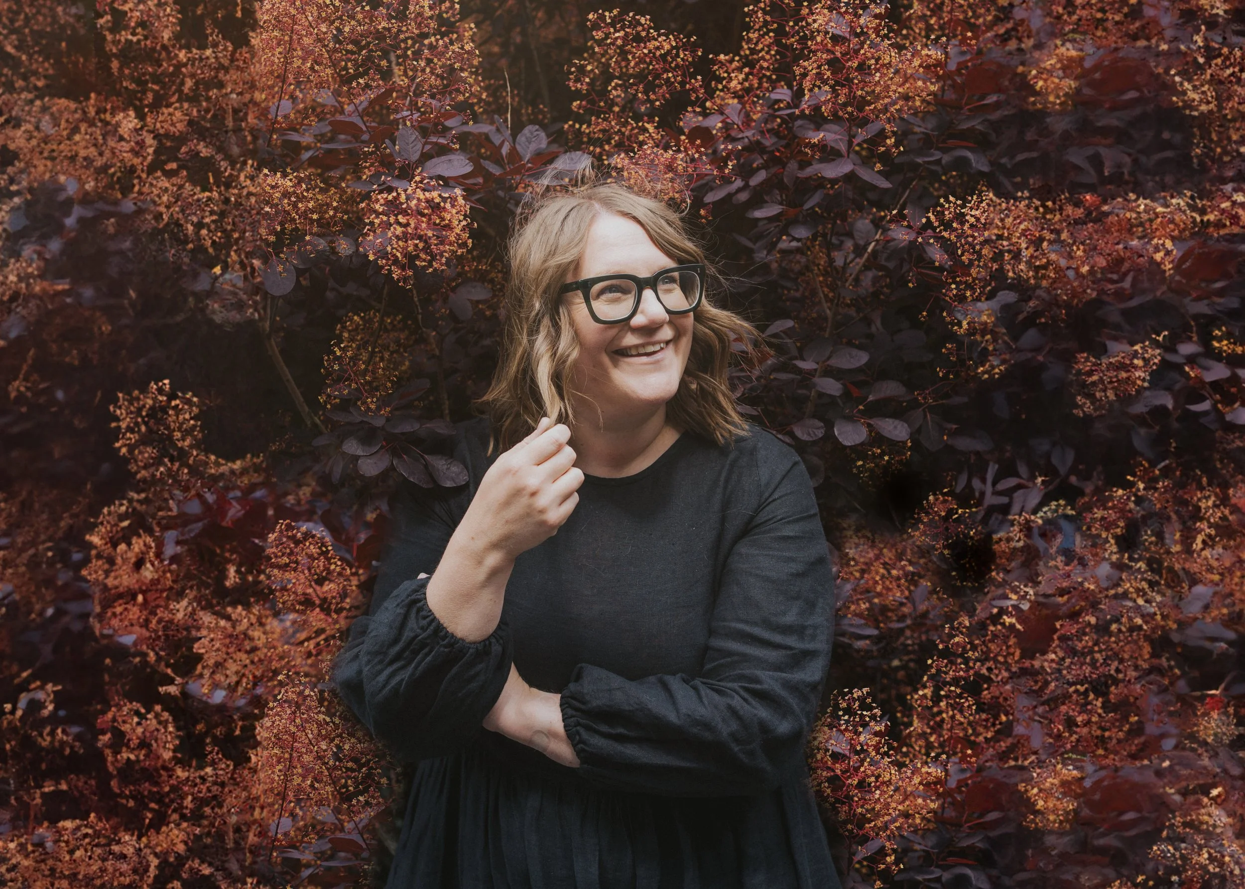 A woman with shoulder-length wavy blonde hair, glasses, and a black dress smiling and standing in front of a dark purple and pink flowering bush.