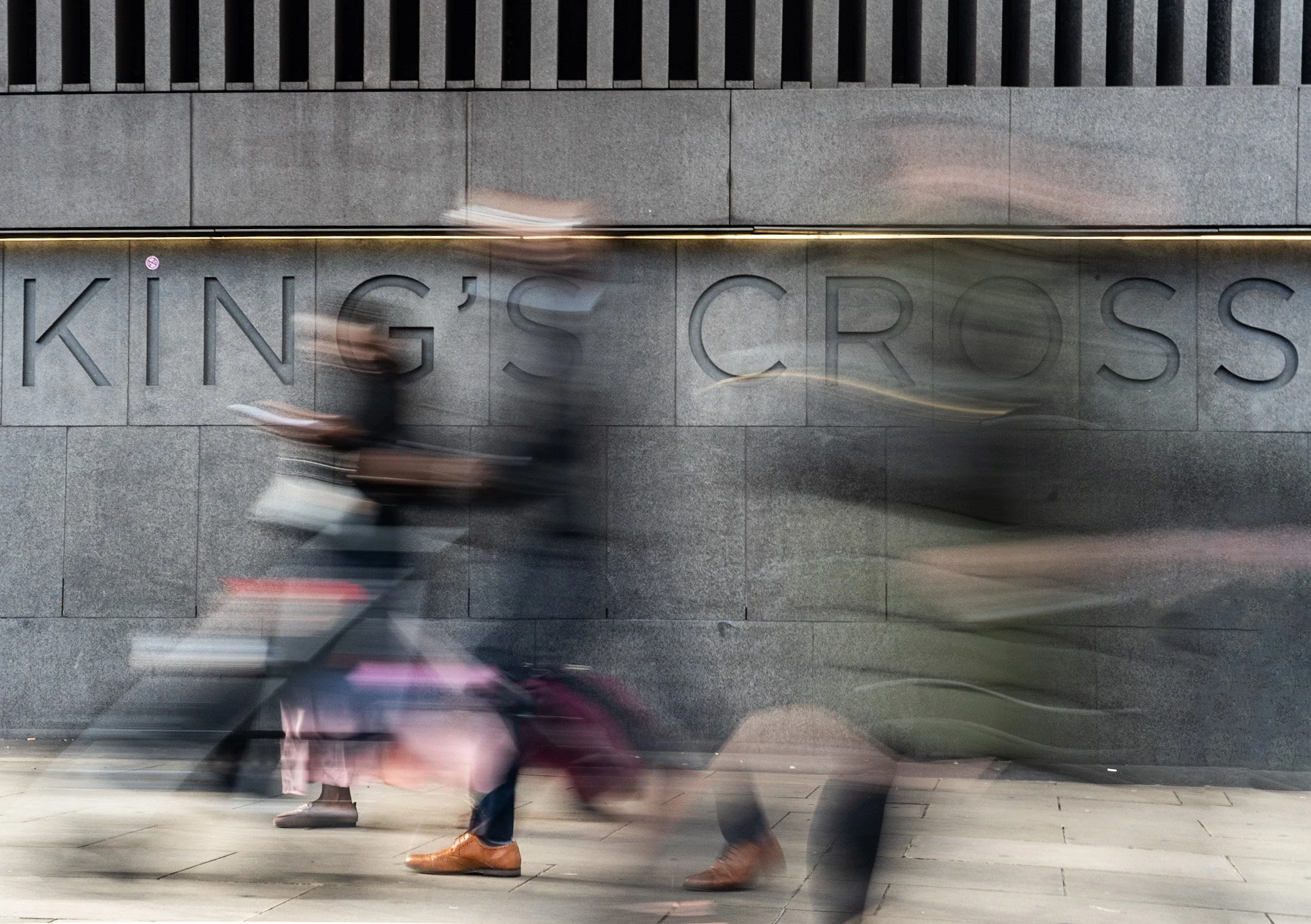 Blurred pedestrians walking past a wall with the words 'KING'S CROSS' engraved in it.
