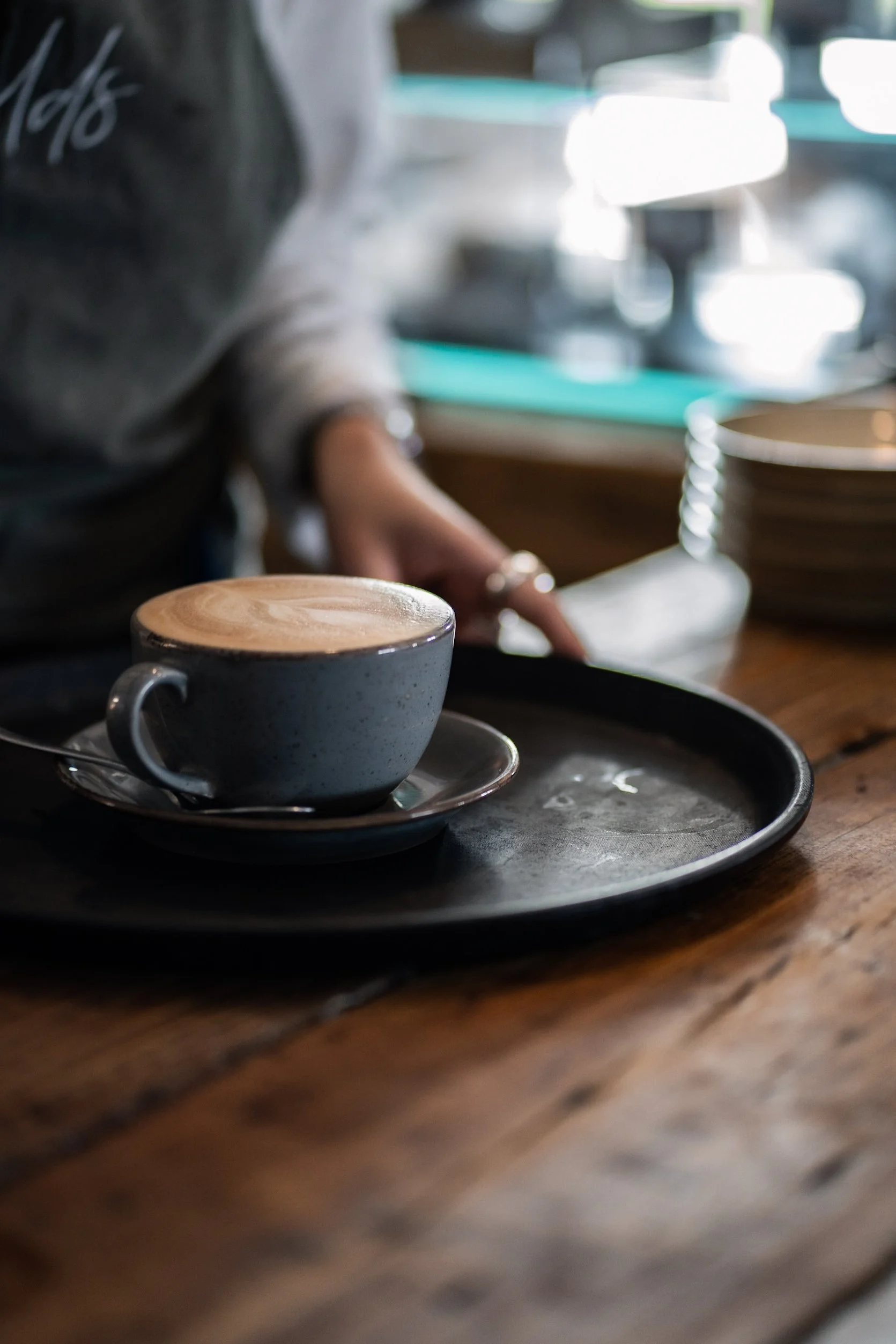 A gray ceramic cup filled with frothy coffee or latte on a black tray, placed on a wooden table at a coffee shop.