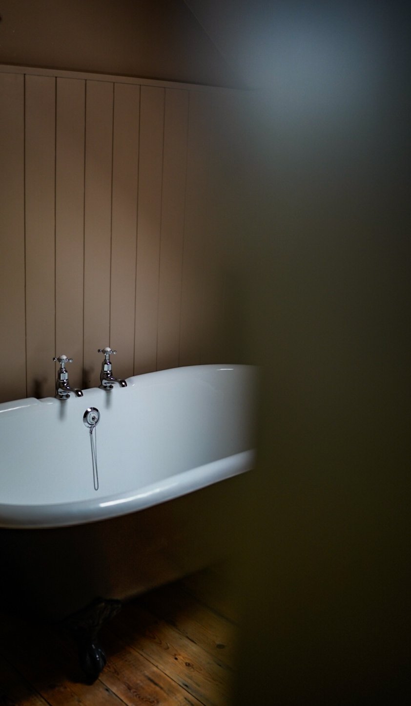 A white side bathtub with three vintage-style faucets, set against a wood-paneled wall in a dimly lit room with hardwood flooring.