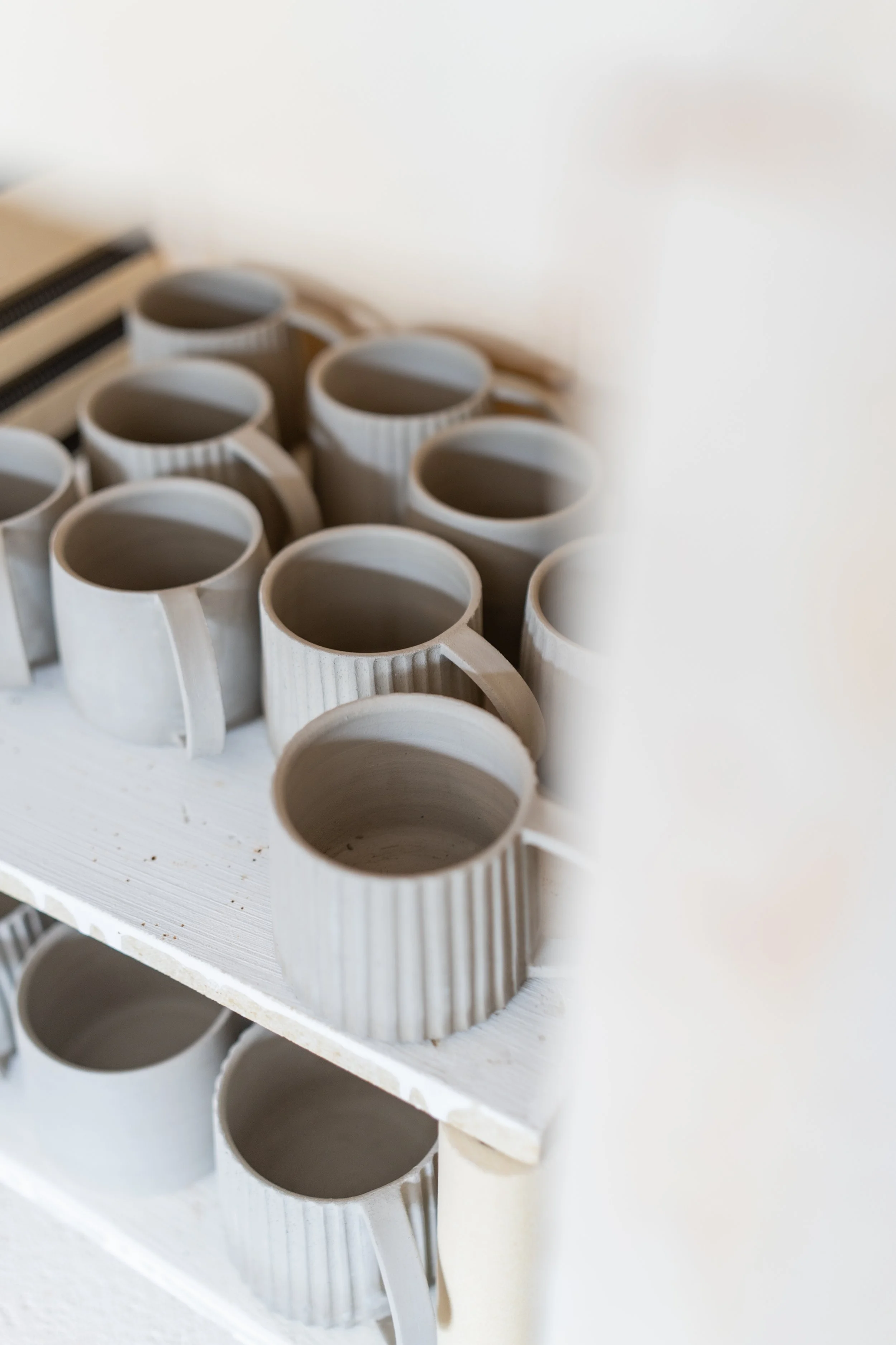 Multiple unfinished ceramic mugs stacked on shelves in a pottery studio.