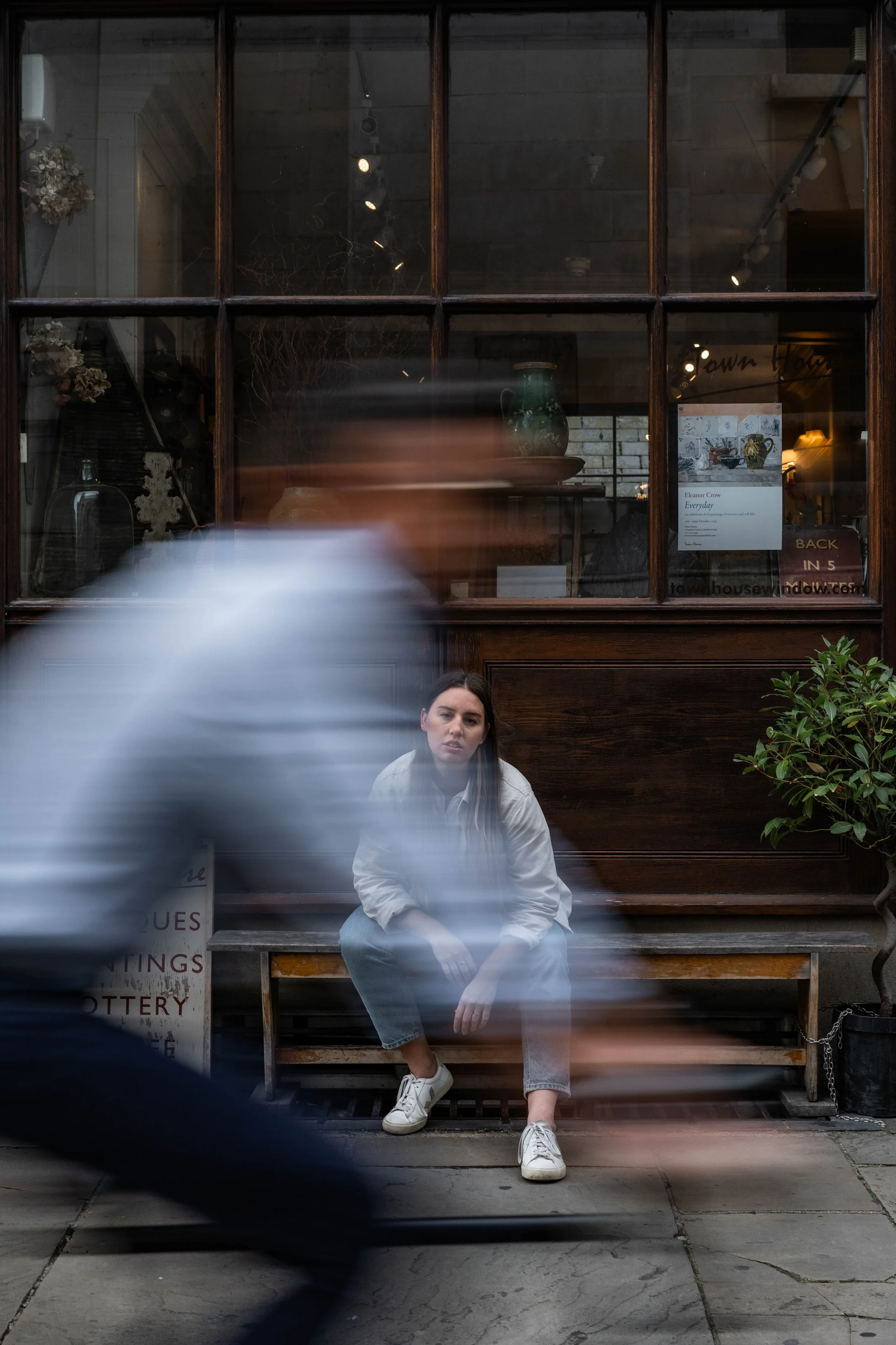 A young woman with long dark hair, wearing a white jacket, gray jeans, and white sneakers, sitting on a bench on a city sidewalk in front of a building with a large window. A blurred person is walking past in front of her.