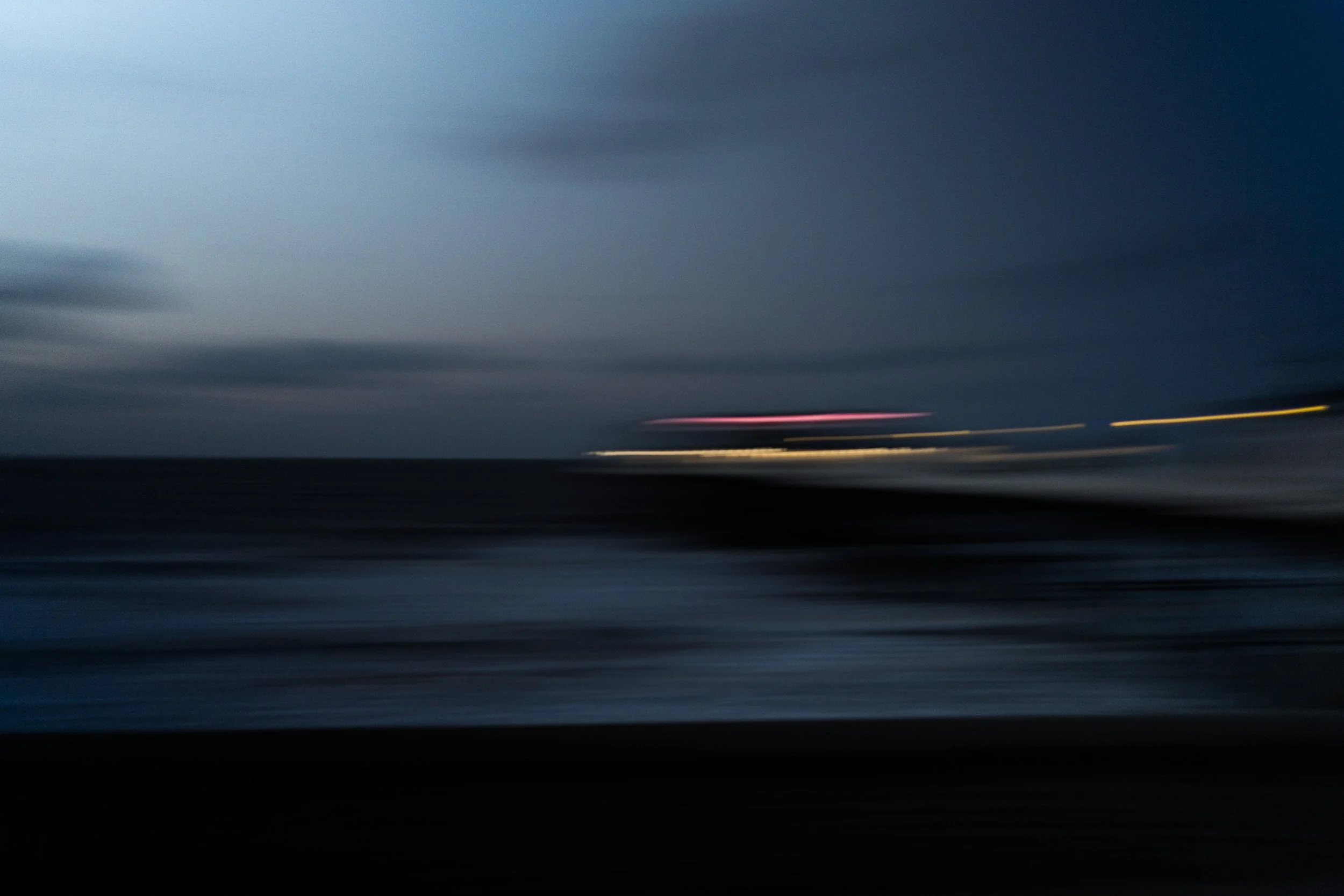 Blurry image of a dark cloudy sky over a body of water with distant lights streaking across the horizon.