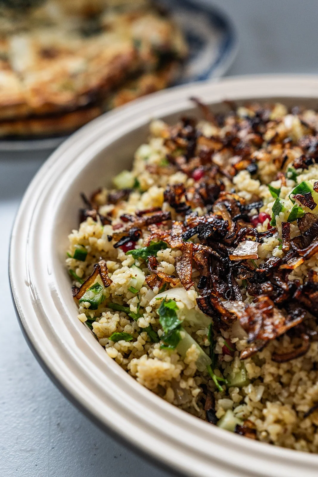 Close-up of a dish in a white bowl with cooked bulgur, chopped vegetables, and caramelized onions, with a baked flatbread in the blurred background.
