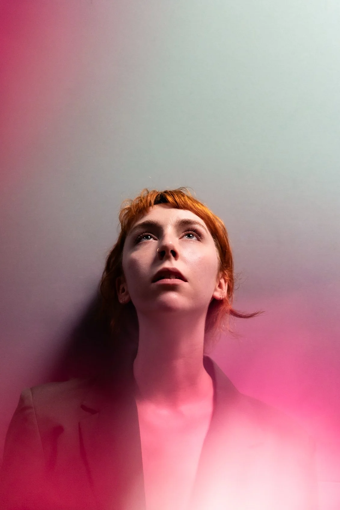 A young woman with short red hair looking upward against a plain background with pink and green light effects.